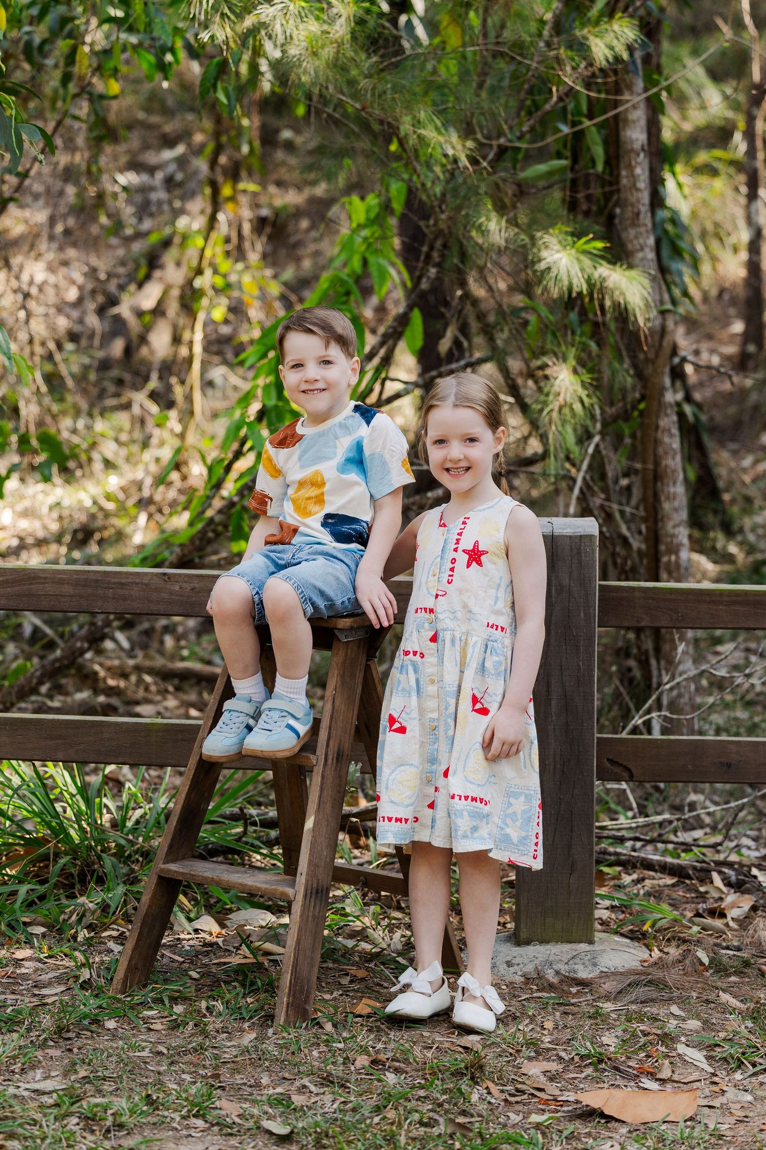 Two children sit and stand by a wooden bench in a lush garden setting with green foliage in the background.