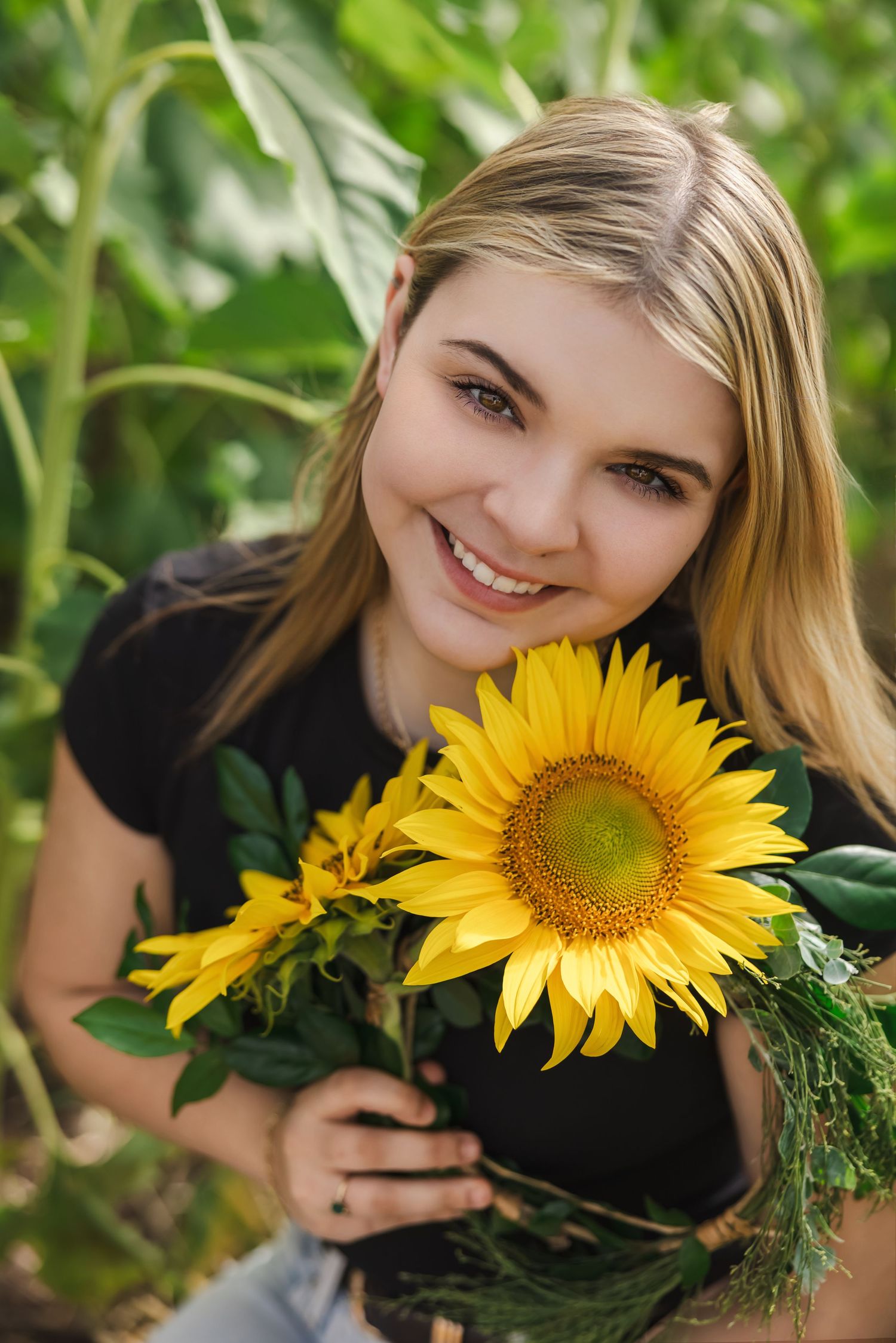 Smiling blonde teenage girl holding a bright yellow sunflower in a lush green garden setting.