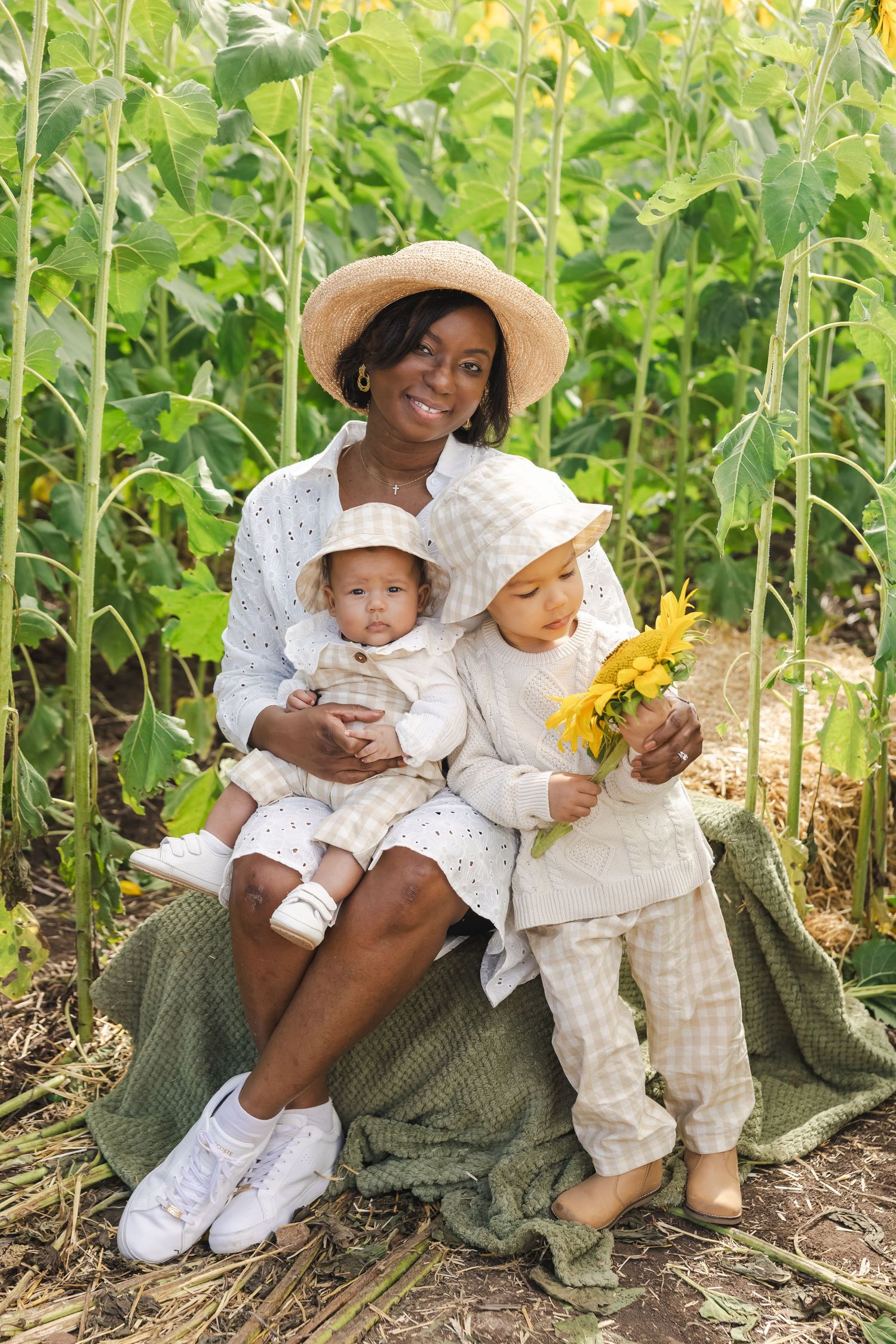 A smiling Black mother in a straw hat sits outdoors holding two young children in neutral outfits near sunflower plants.