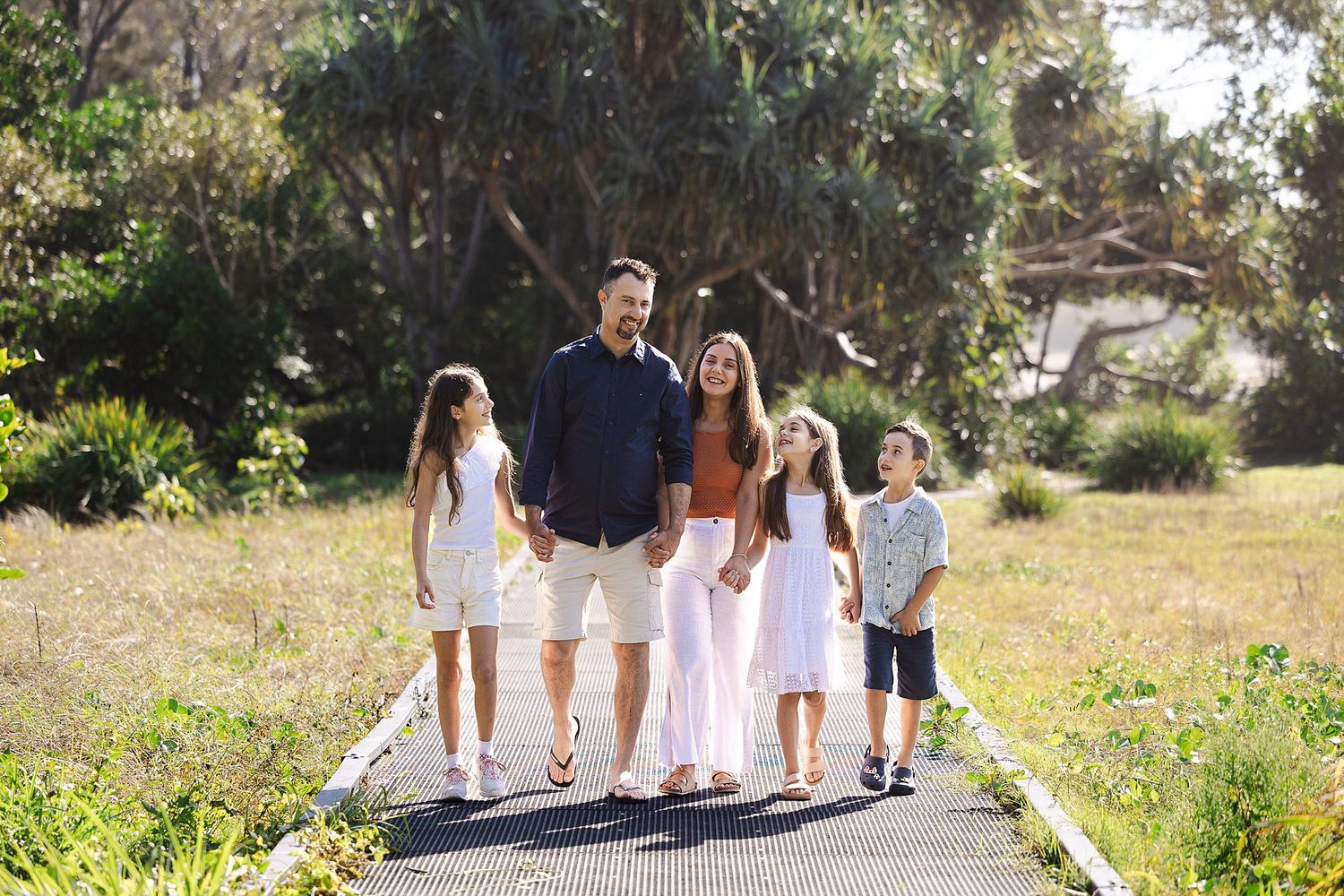 Family of five walking together on a paved pathway surrounded by lush greenery on a sunny day.