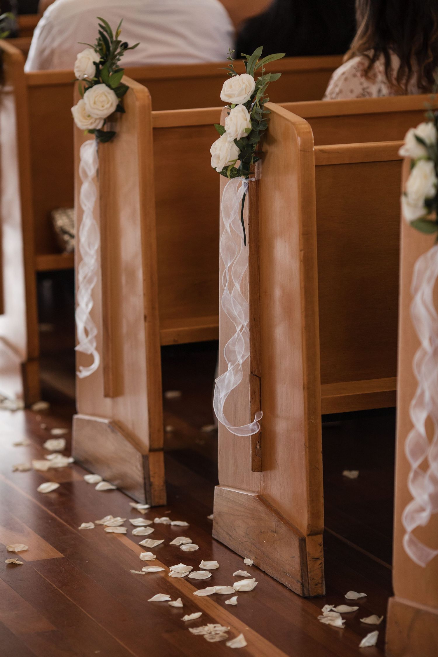 Church wedding aisle with wooden pews decorated with white roses, ribbon, and scattered flower petals on hardwood floor.