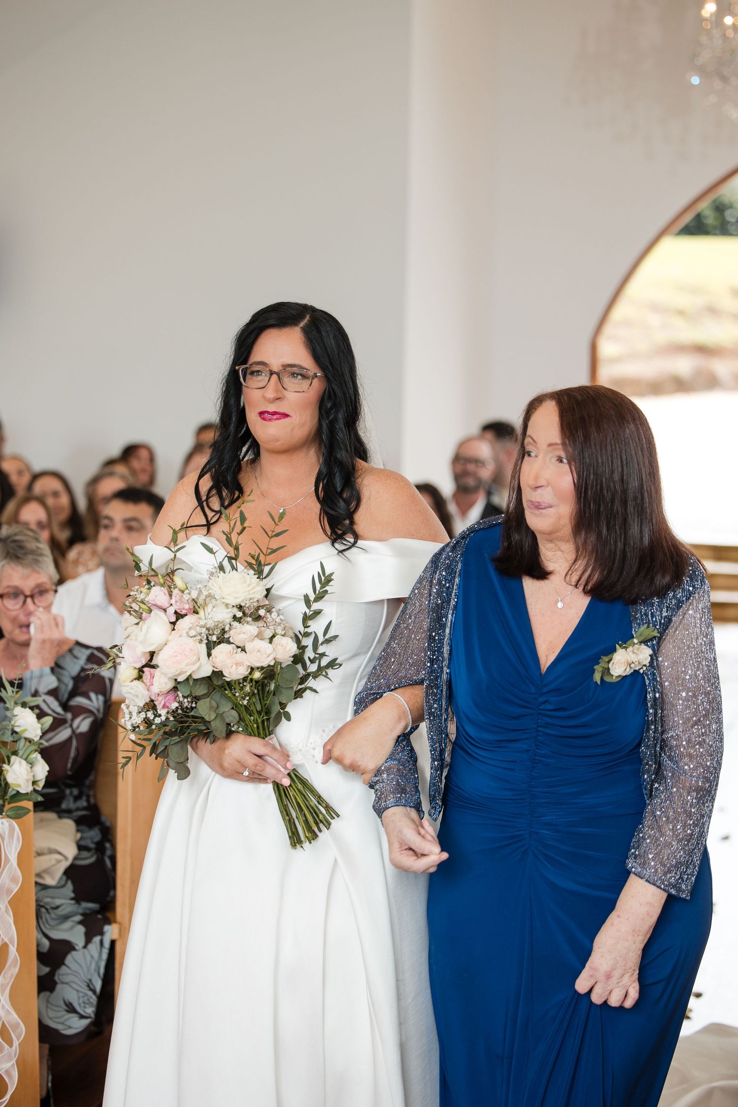 Bride in white off-shoulder gown holding floral bouquet walks down aisle with woman in blue sequin dress.