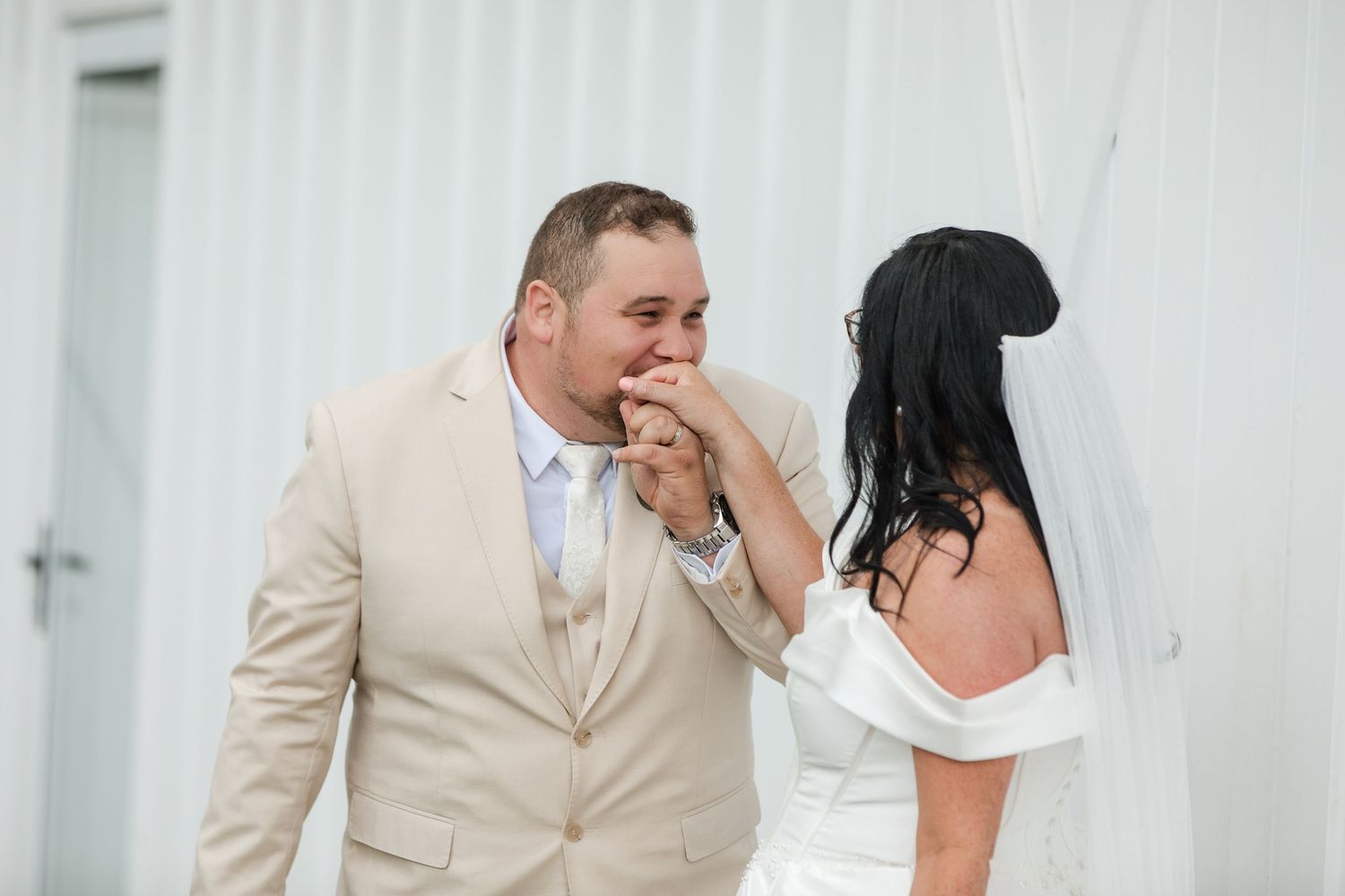 Groom in beige suit kissing bride's hand during outdoor wedding ceremony, bride wearing white off-shoulder gown and veil.