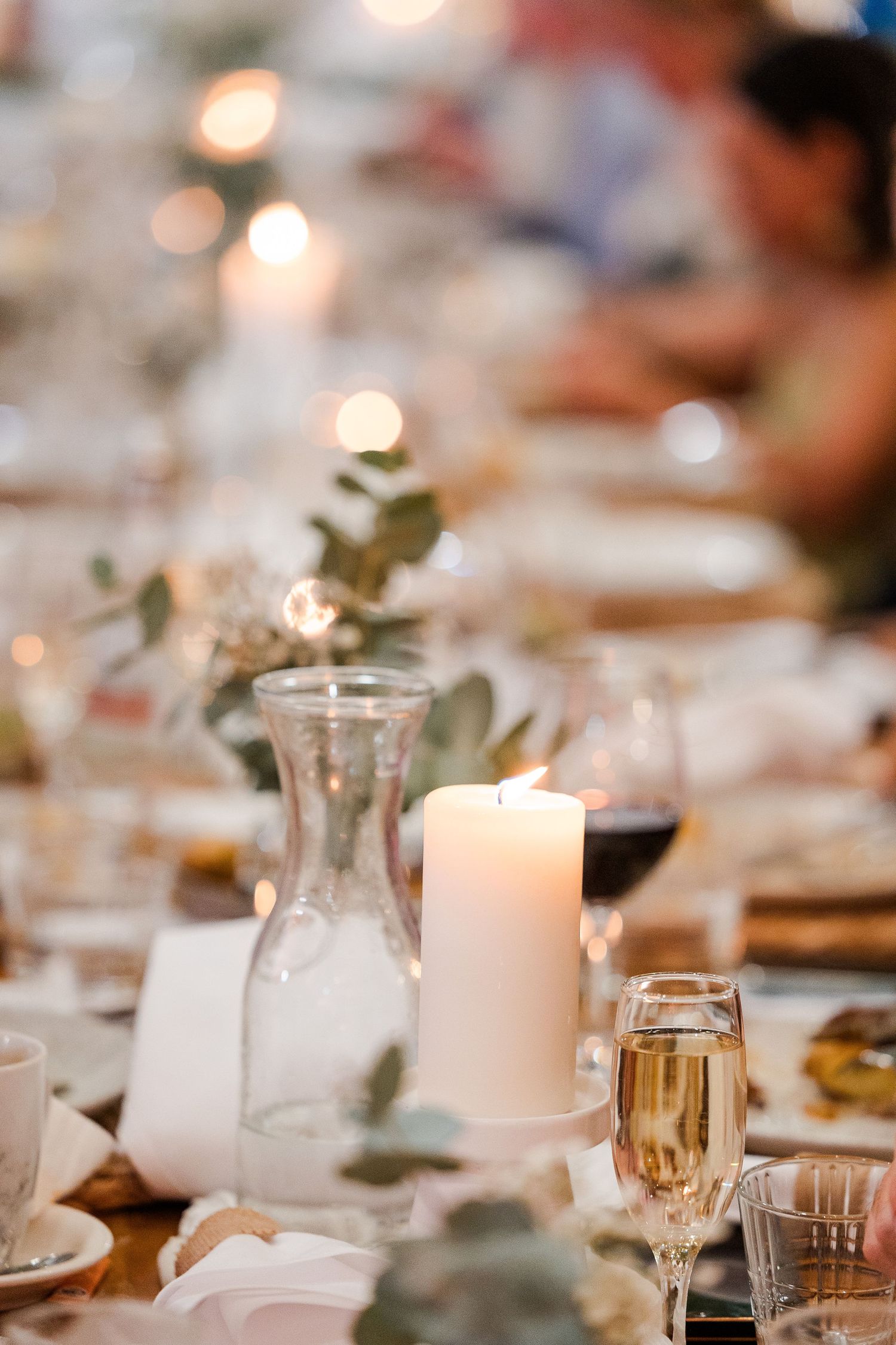 Elegant wedding reception table with lit pillar candle, champagne flute, eucalyptus greenery, and glass carafe centerpiece.
