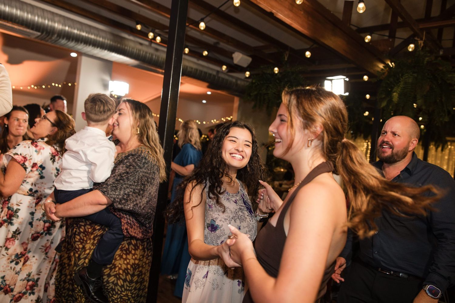 Guests laughing and socializing at an indoor wedding reception with warm string lights in the background.