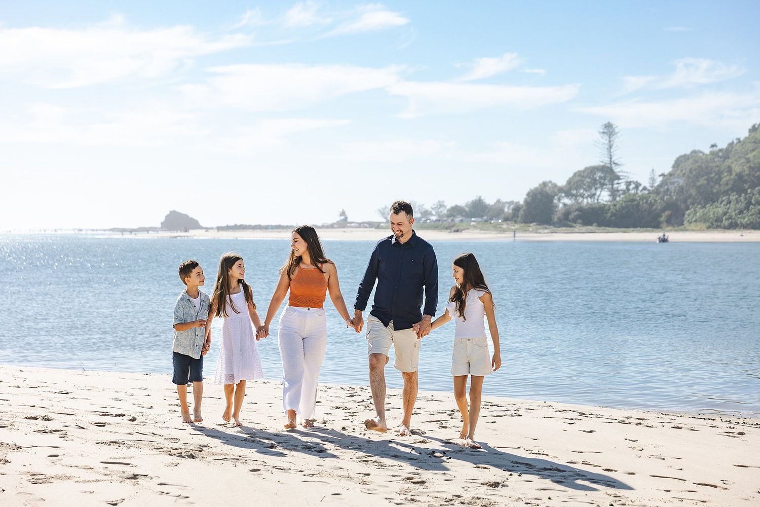 Group in summer clothing walking together along a tranquil beach shoreline with coastal scenery and blue sky in background.