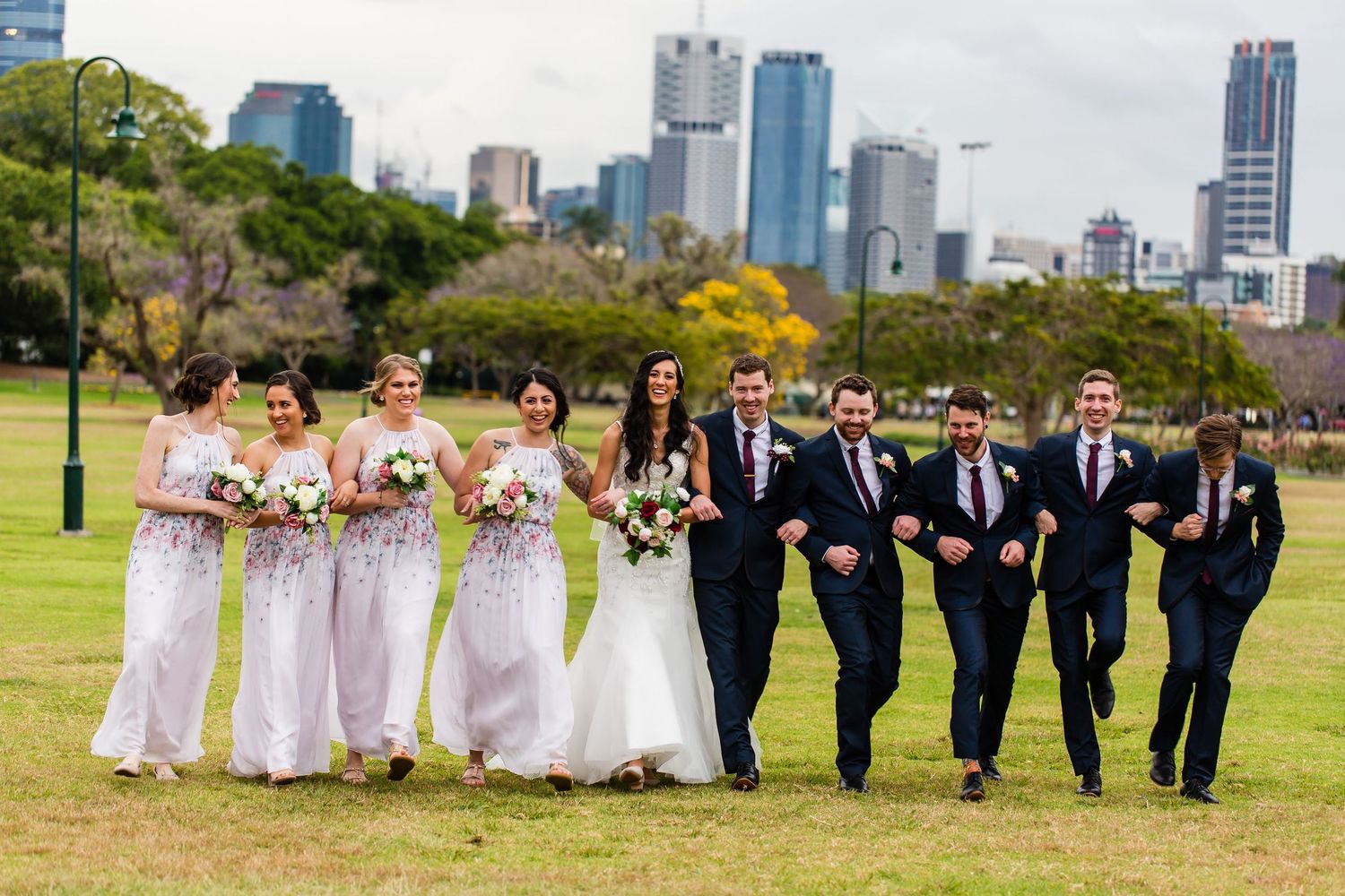 Wedding party walking together on grassy lawn with city skyline in background during outdoor celebration.
