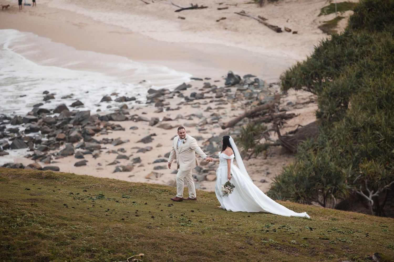 Bride and groom walk hand-in-hand on a scenic coastal hillside with sandy beach and ocean waves in the background.