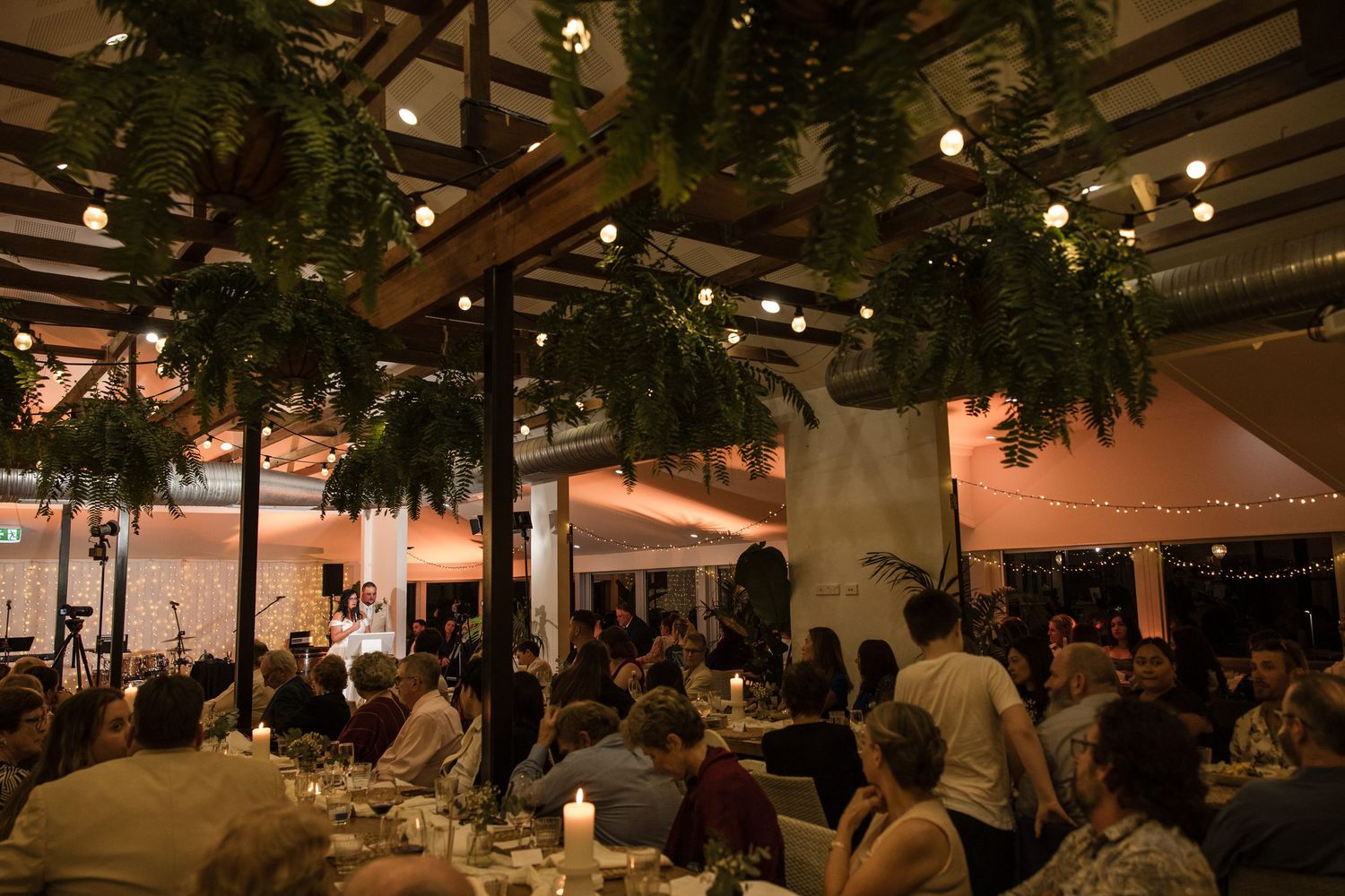 Elegant outdoor dinner event with string lights, lush fern decor, and guests dining under a pergola at night.