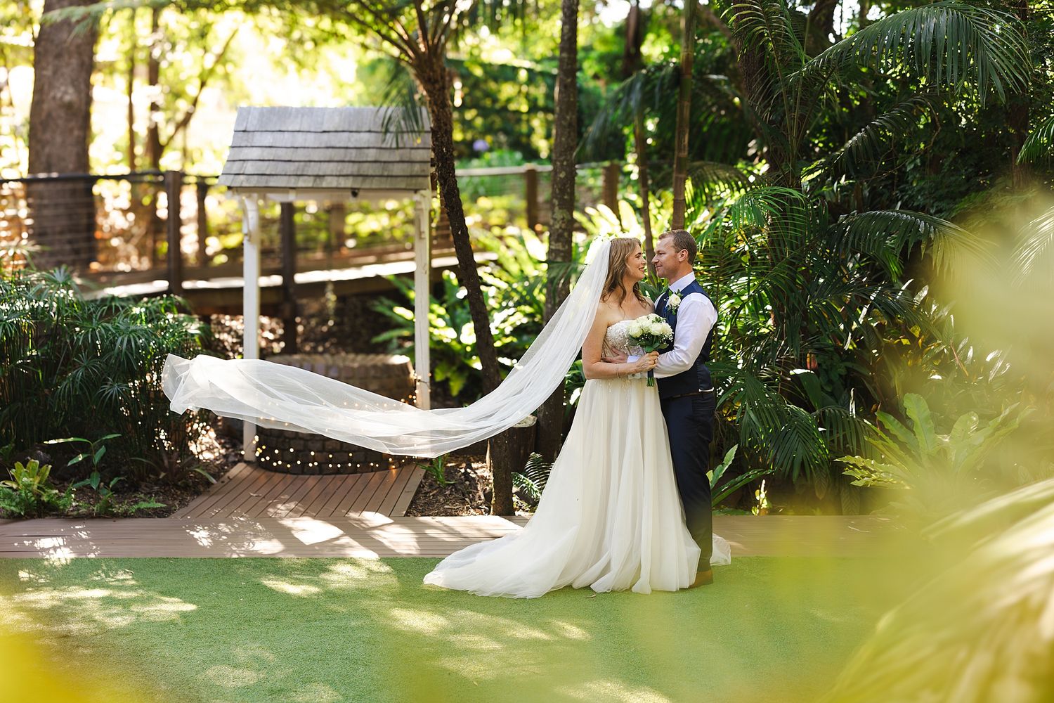 A couple embraces in a lush garden setting with natural sunlight filtering through the trees.