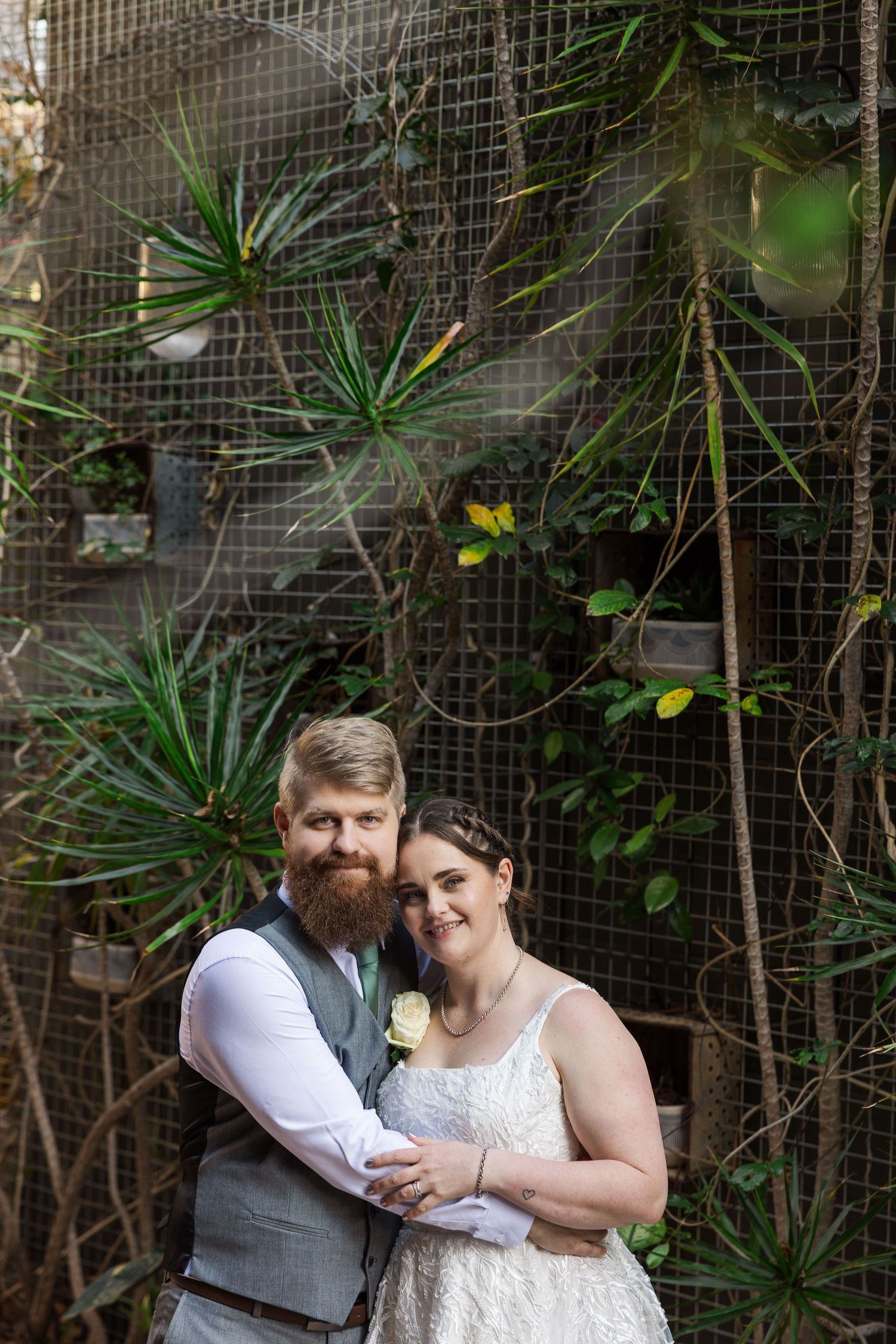 Smiling bride and groom posing together in front of a lush green plant wall on their wedding day.