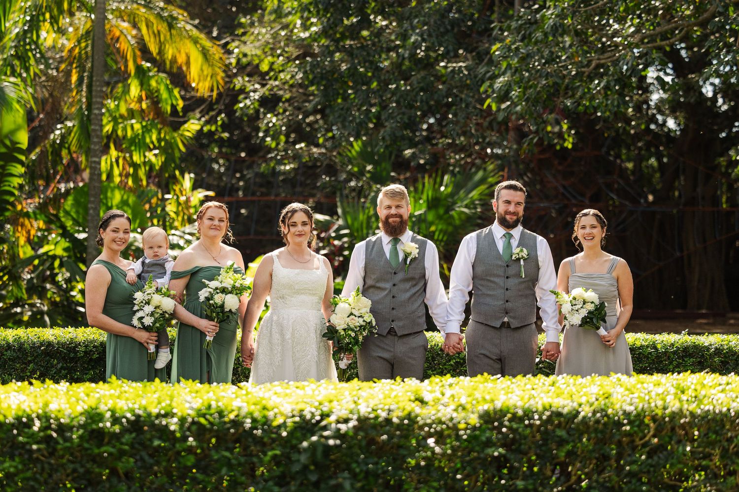 Wedding party poses outdoors in lush garden, bridesmaids in sage green dresses, groomsmen in grey suits holding white bouquets.