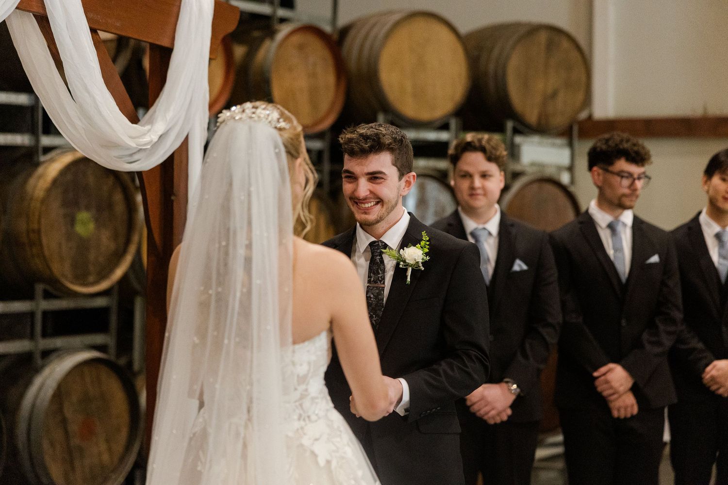 Groom smiling at bride during wedding ceremony in rustic winery with wooden barrels and groomsmen standing behind.