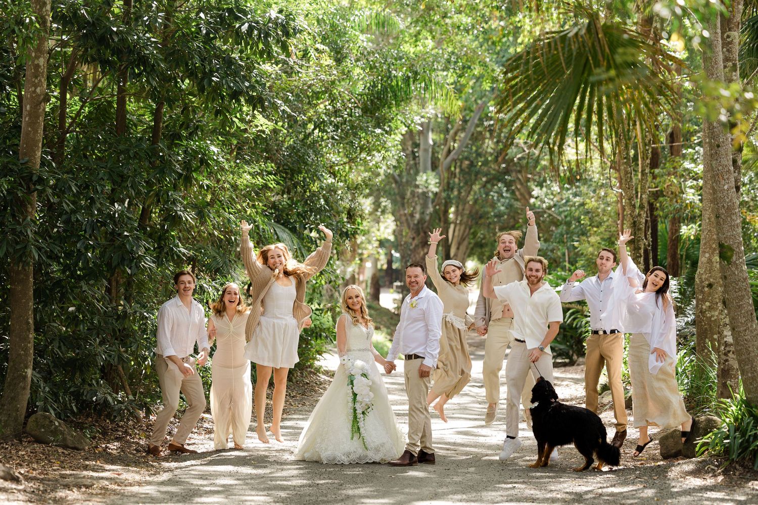 Joyful wedding party celebrating outdoors on a tree-lined path with a black dog, all dressed in white and beige attire.
