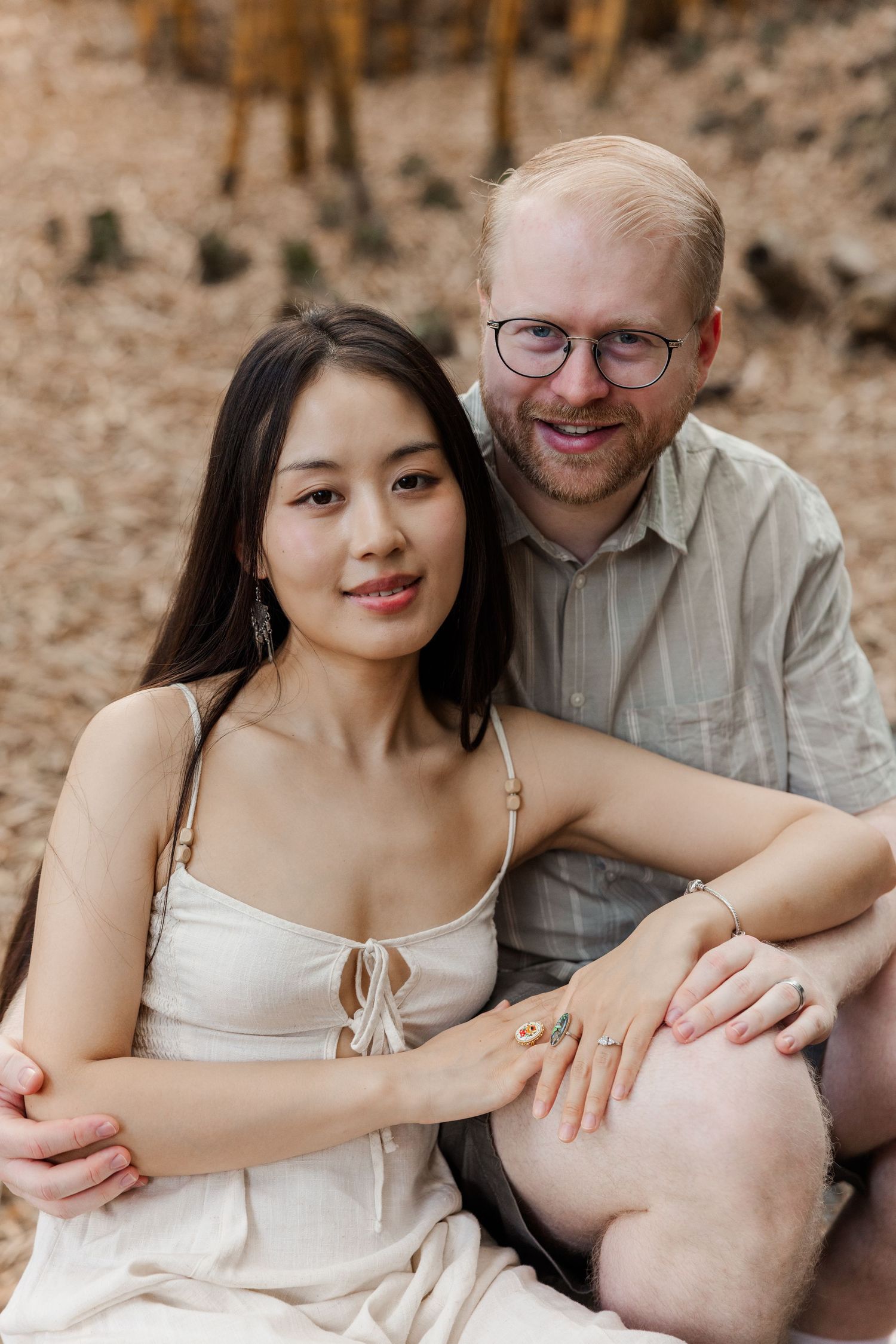 Couple sitting close together on ground during outdoor photo session with natural backdrop.