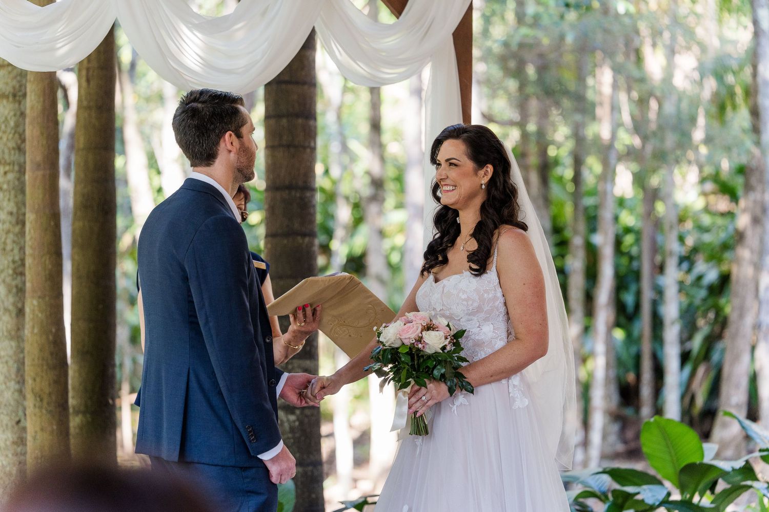 A romantic wedding ceremony scene under tall palm trees in a lush tropical garden setting.