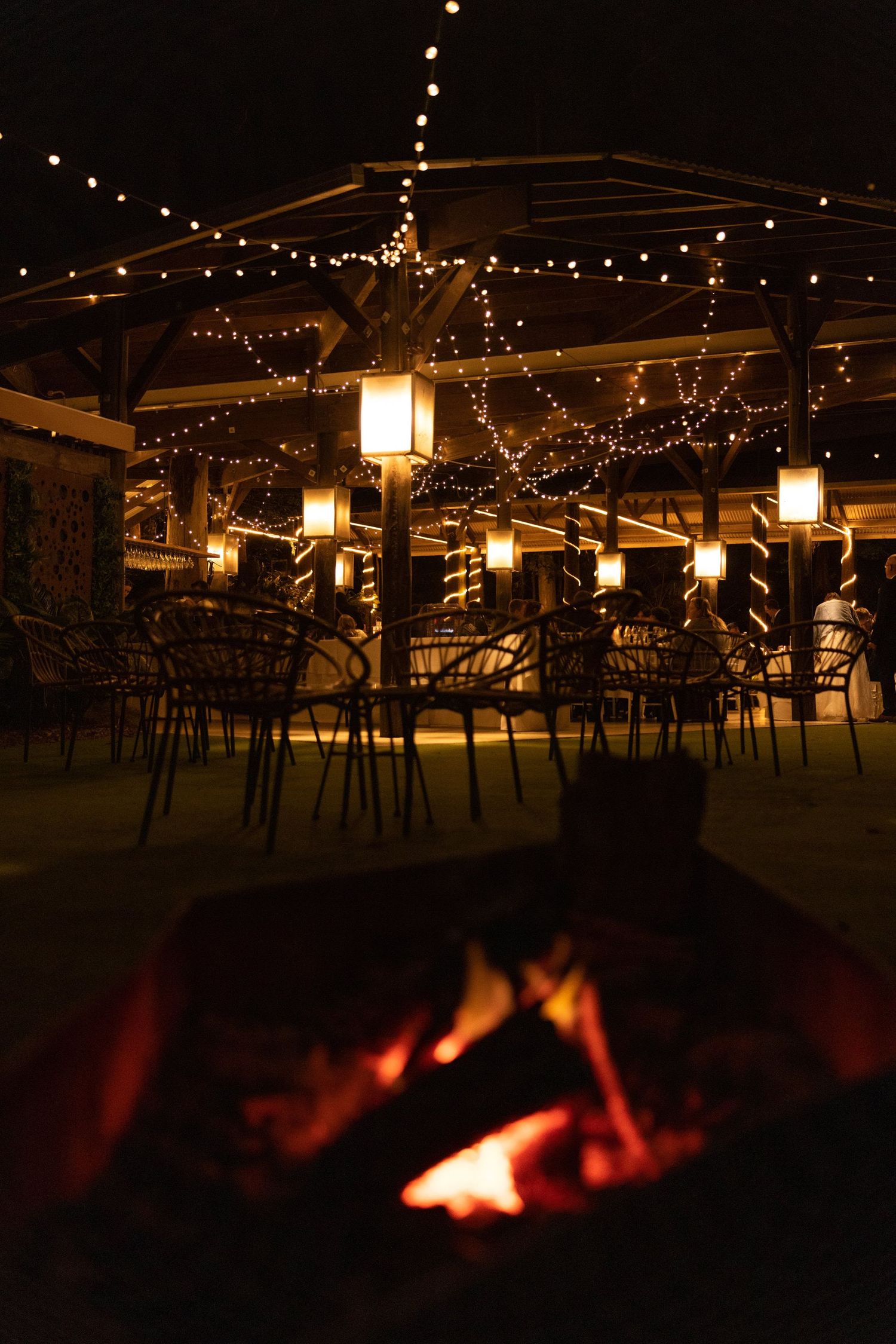 String lights illuminate an outdoor patio dining area at night with a warm glowing fire pit in the foreground.