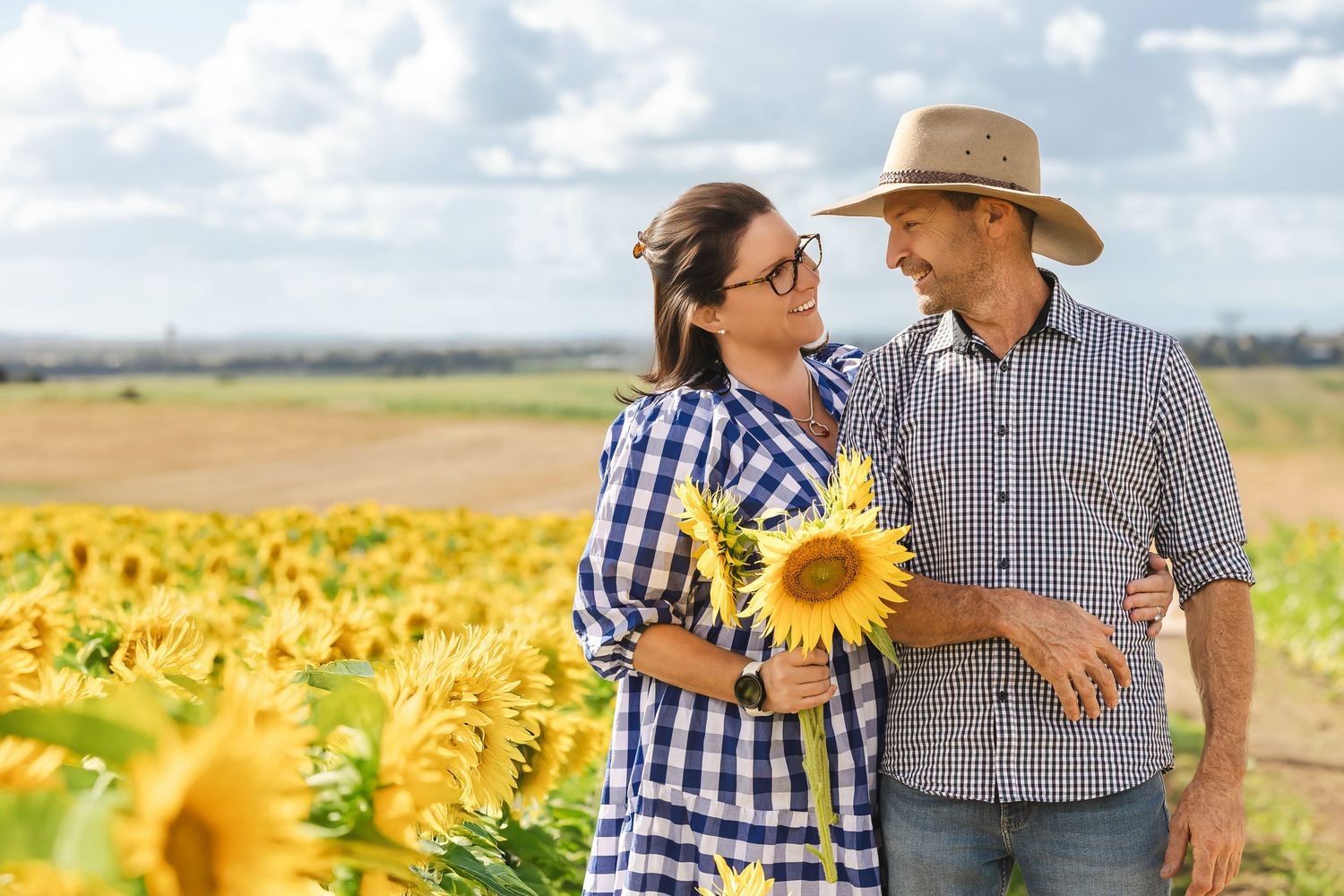 Smiling couple in plaid shirts holding sunflowers while walking through a bright sunflower field.