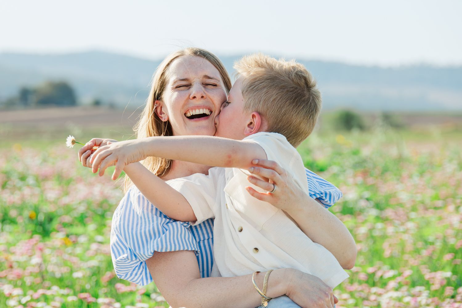 A laughing woman holds a young boy in a sunny flower field as he kisses her cheek and hands her a flower.