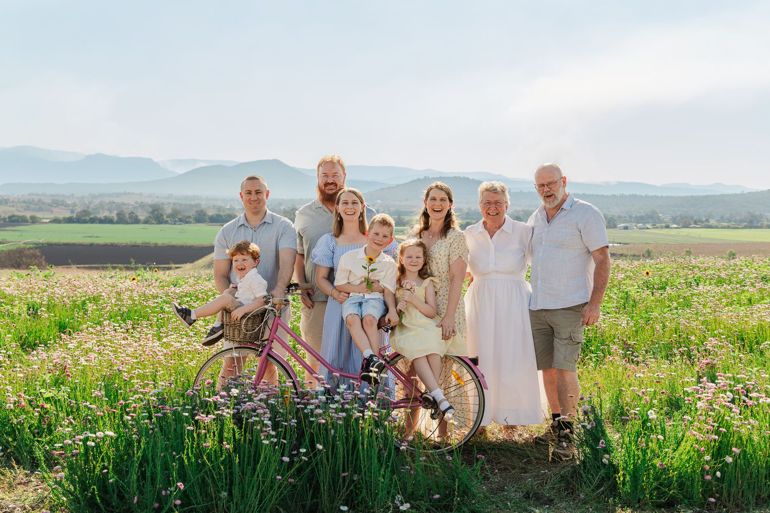 Large family posing together in a sunny green field with bicycles and mountain views in the background.