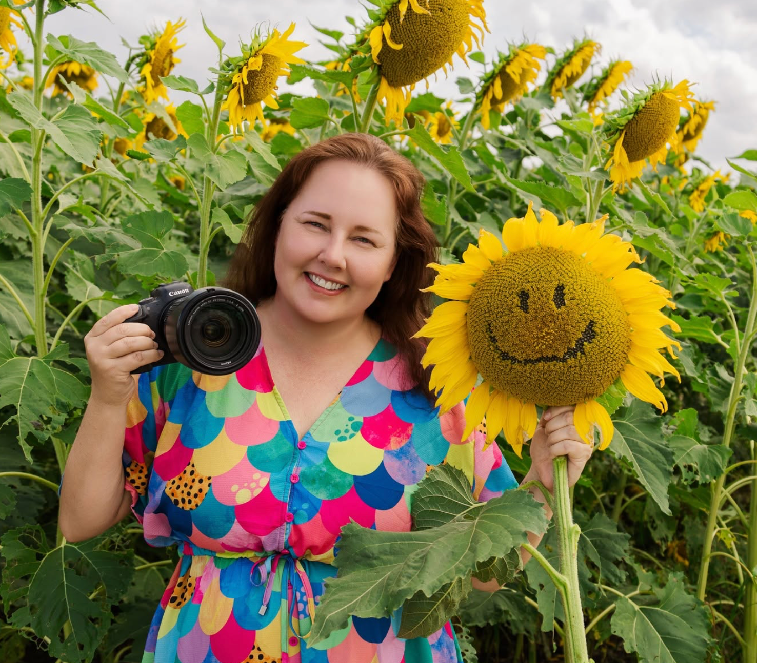 Woman in colorful dress holds camera and smiley face sunflower in a sunny sunflower field.