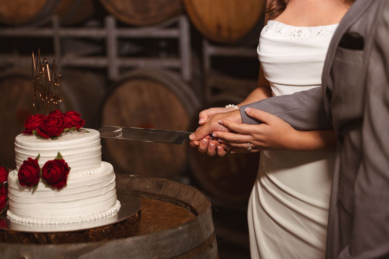 Bride and groom cutting a white wedding cake with red roses on a wooden barrel in a rustic winery.