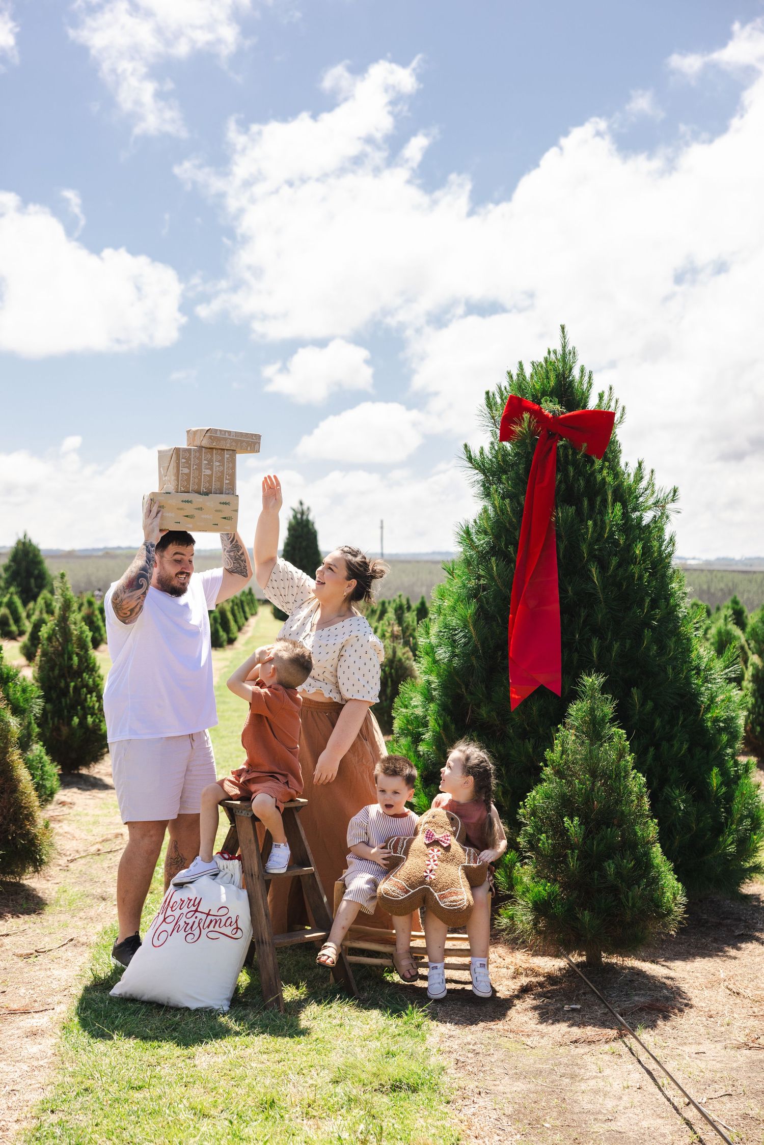 A family takes Christmas photos together at a Christmas tree farm on a sunny day with evergreen trees in the background.