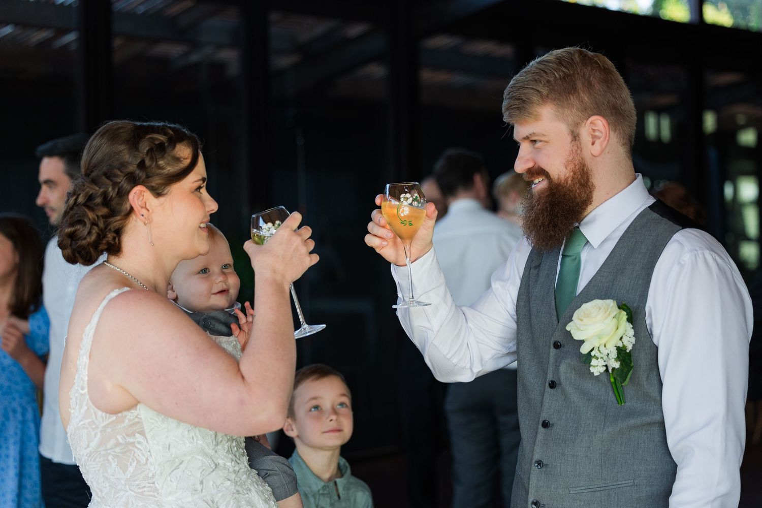 Bride and groom toasting with drinks at wedding reception, groom wearing gray vest with green tie and white boutonniere.