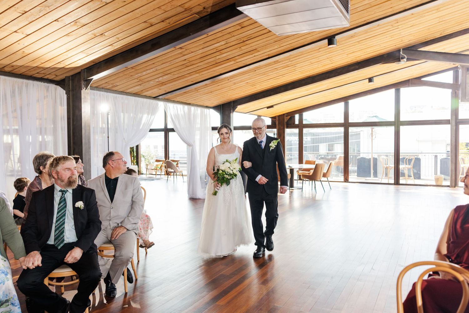 Bride walks down the aisle with her father in an elegant wood-paneled wedding venue with large windows.
