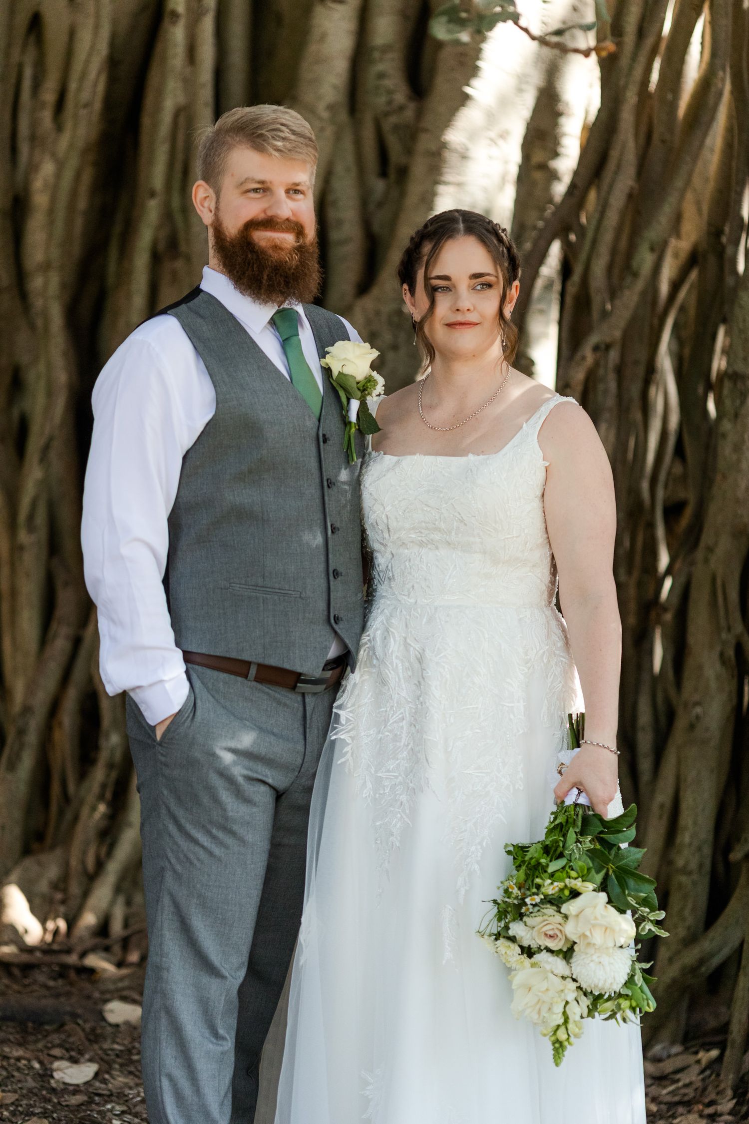 Bride and groom posing together in front of banyan trees, bride holding white floral bouquet in wedding dress.