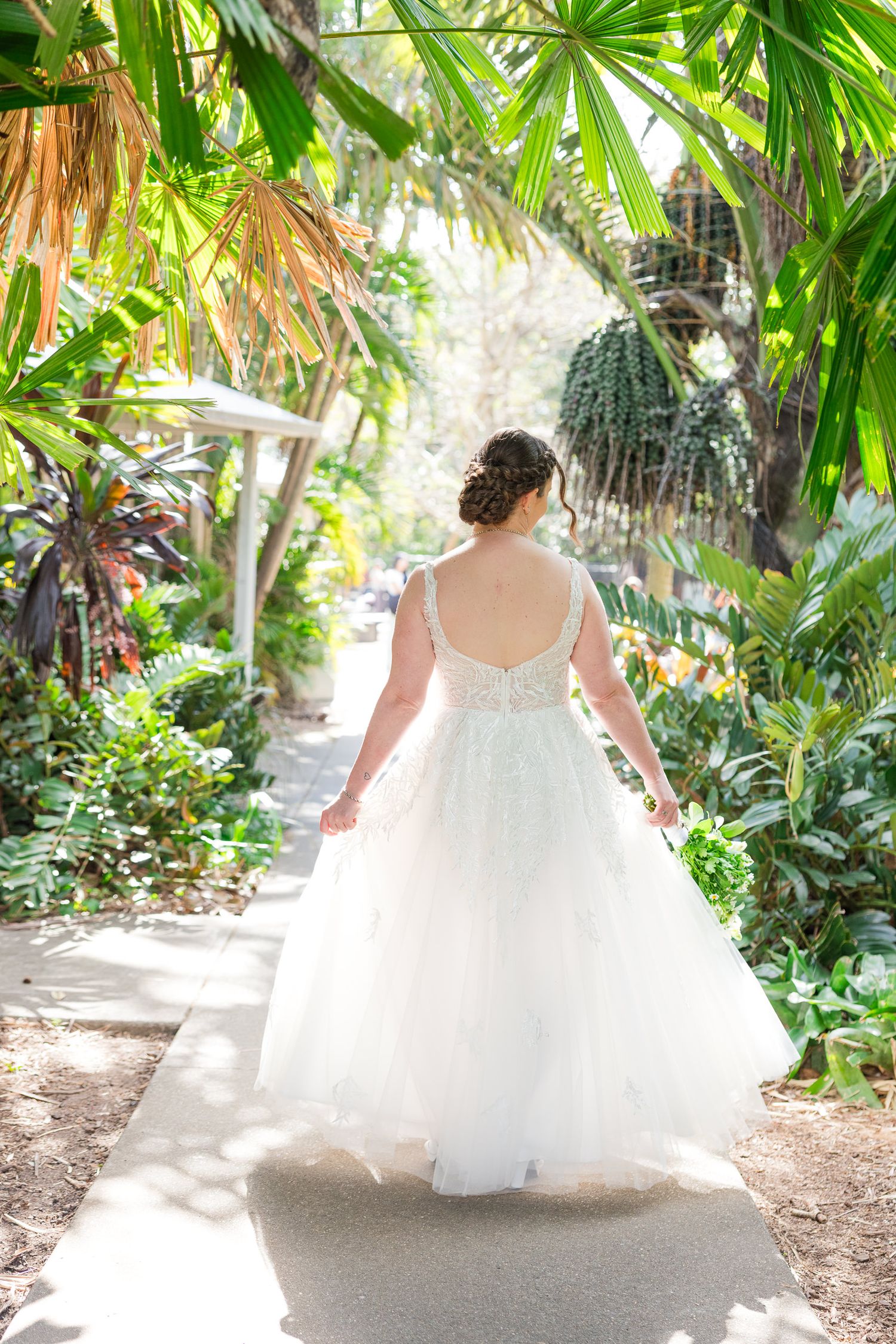 Bride in white lace ballgown walks away down tropical garden path, showing elegant open back and tulle skirt.