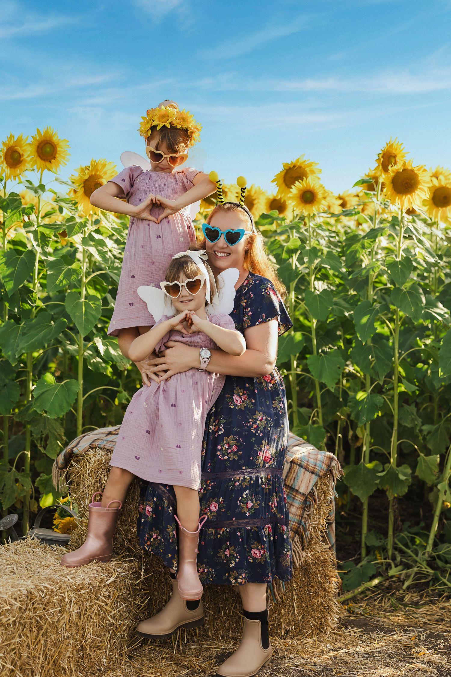 Mother and two daughters wearing sunglasses pose on a hay bale in a sunny sunflower field, girls in matching pink dresses.