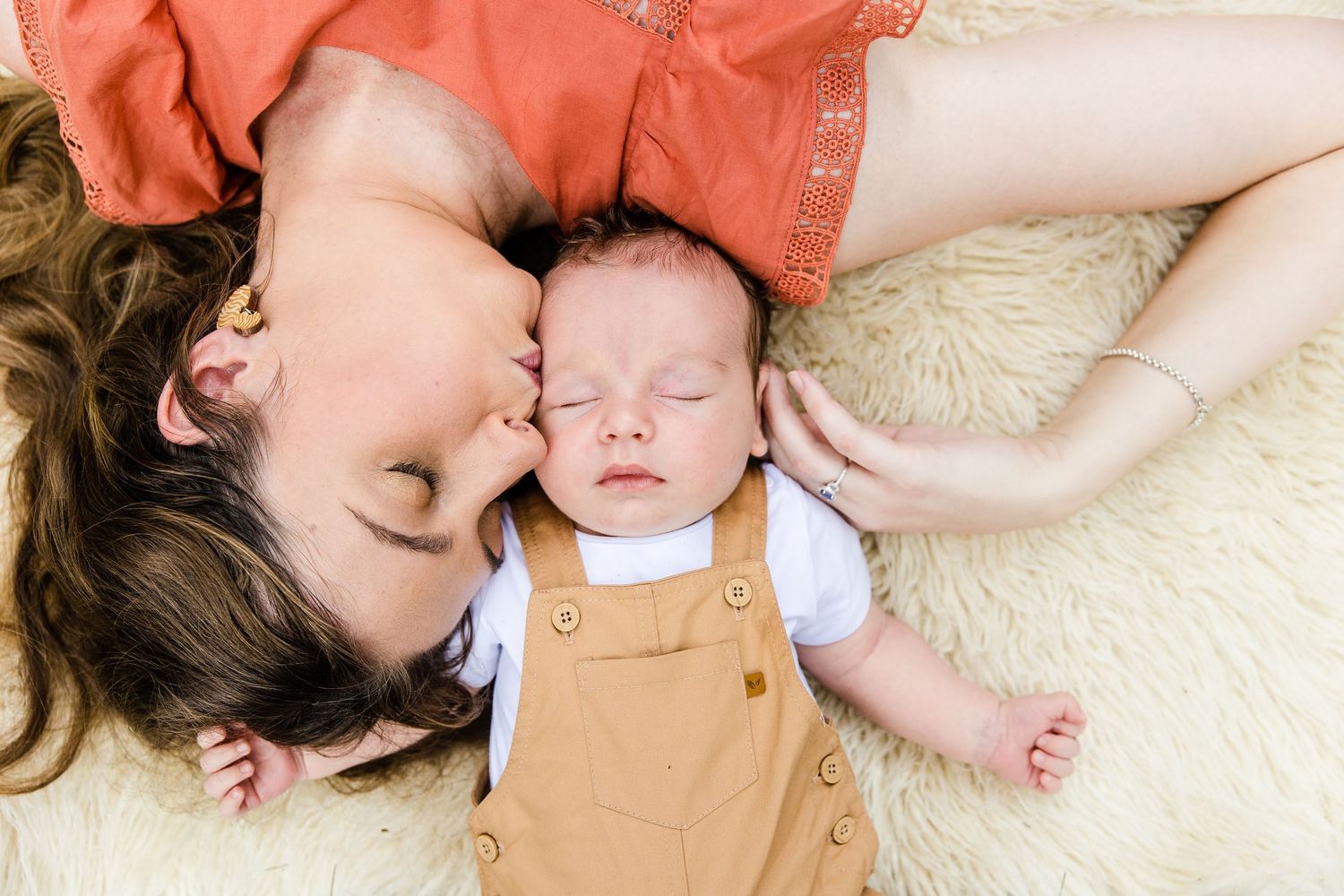 Adult and sleeping baby in tan overalls lying together on plush cream carpet.