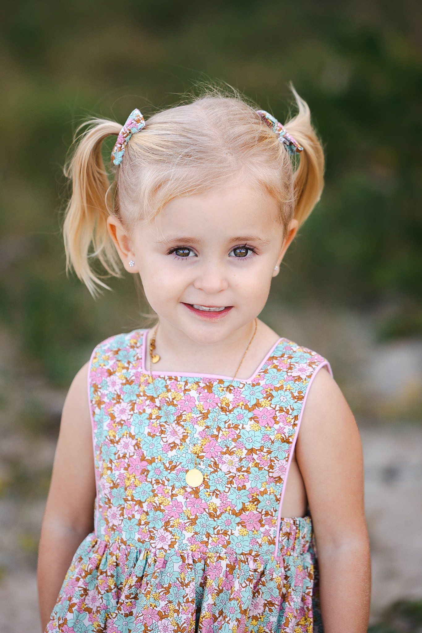 Young child in floral sundress with blonde pigtails smiling at camera in outdoor portrait setting.