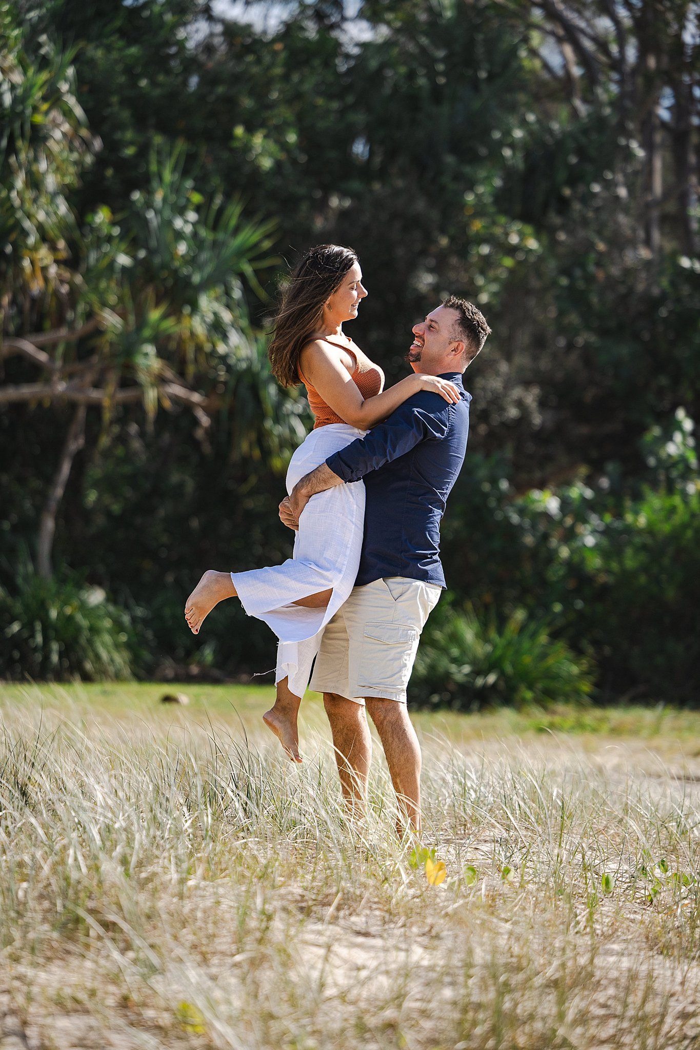 A joyful couple embraces in a romantic pose amidst tall grass and lush greenery on a sunny day.