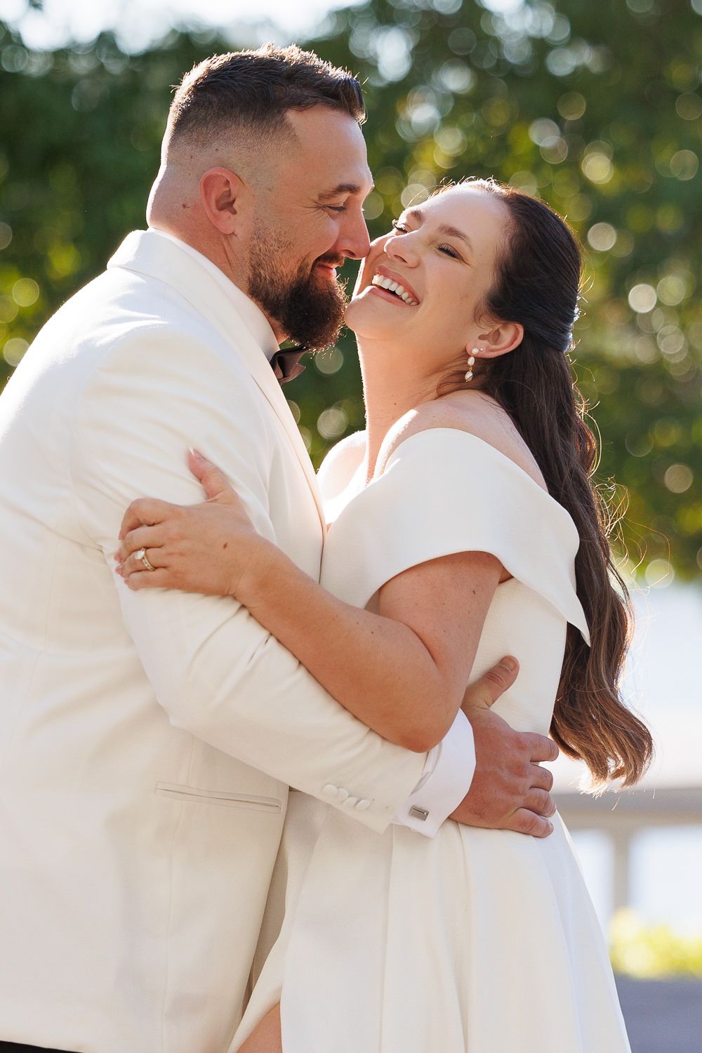 A joyful couple in matching white outfits share an intimate embrace and laugh together outdoors on their wedding day.