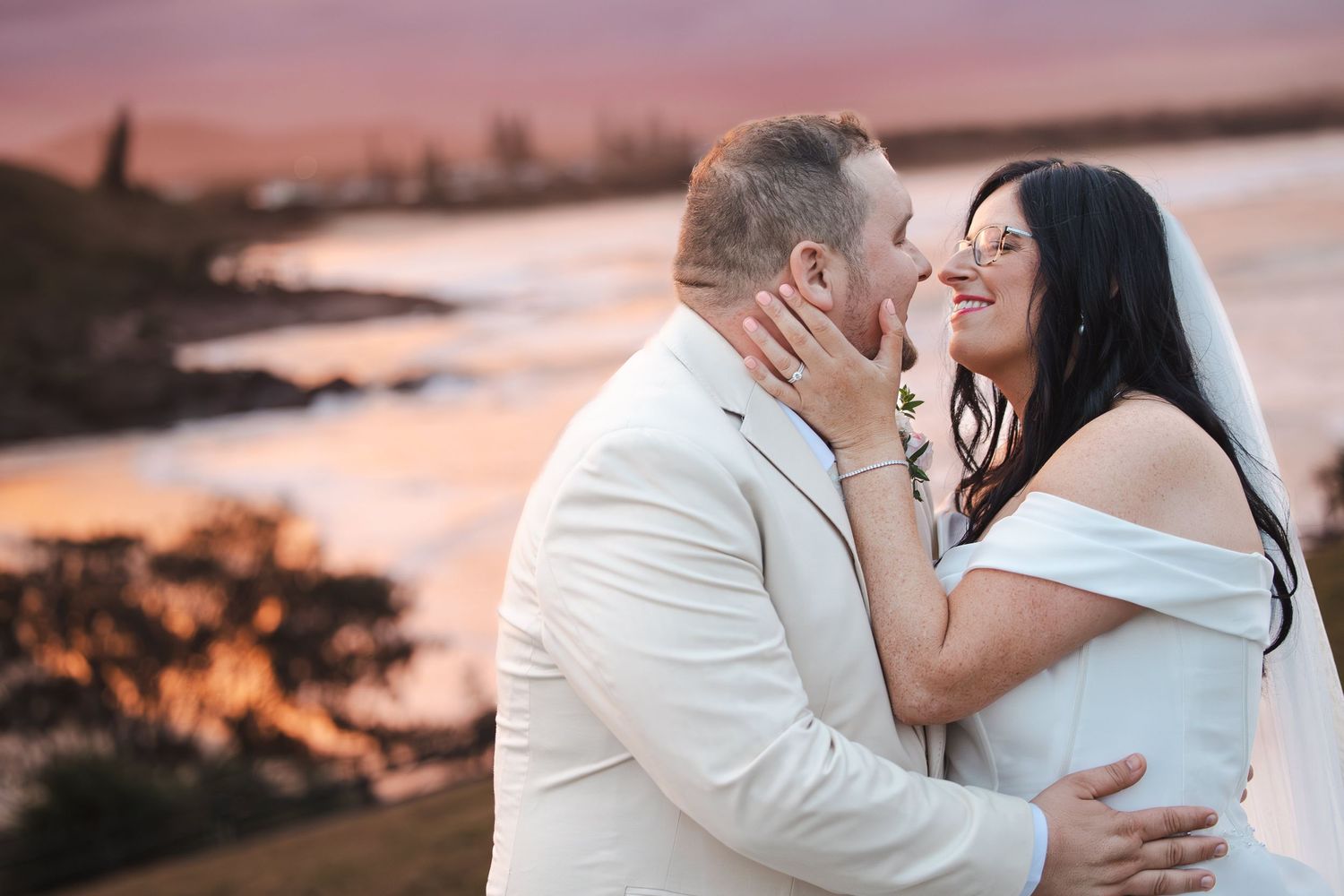 Bride and groom share a romantic moment at sunset by the ocean, bride in white off-shoulder gown with veil.