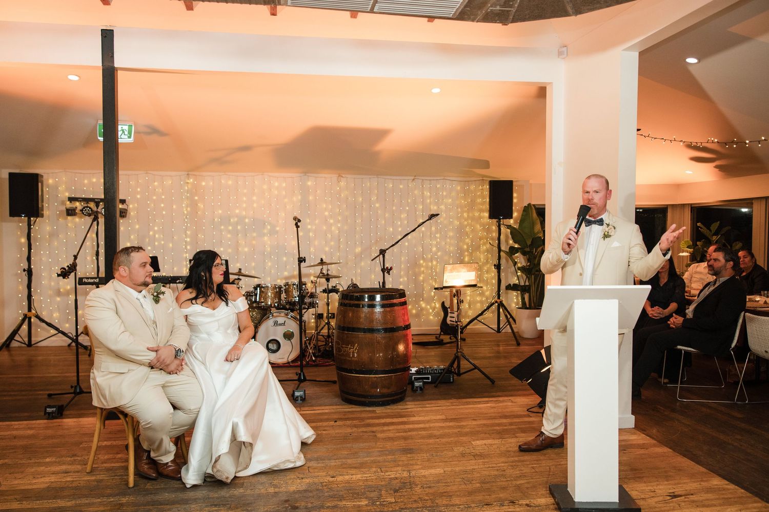 Wedding reception speech at elegant venue with fairy lights, groom at podium addressing seated bride and groom couple.