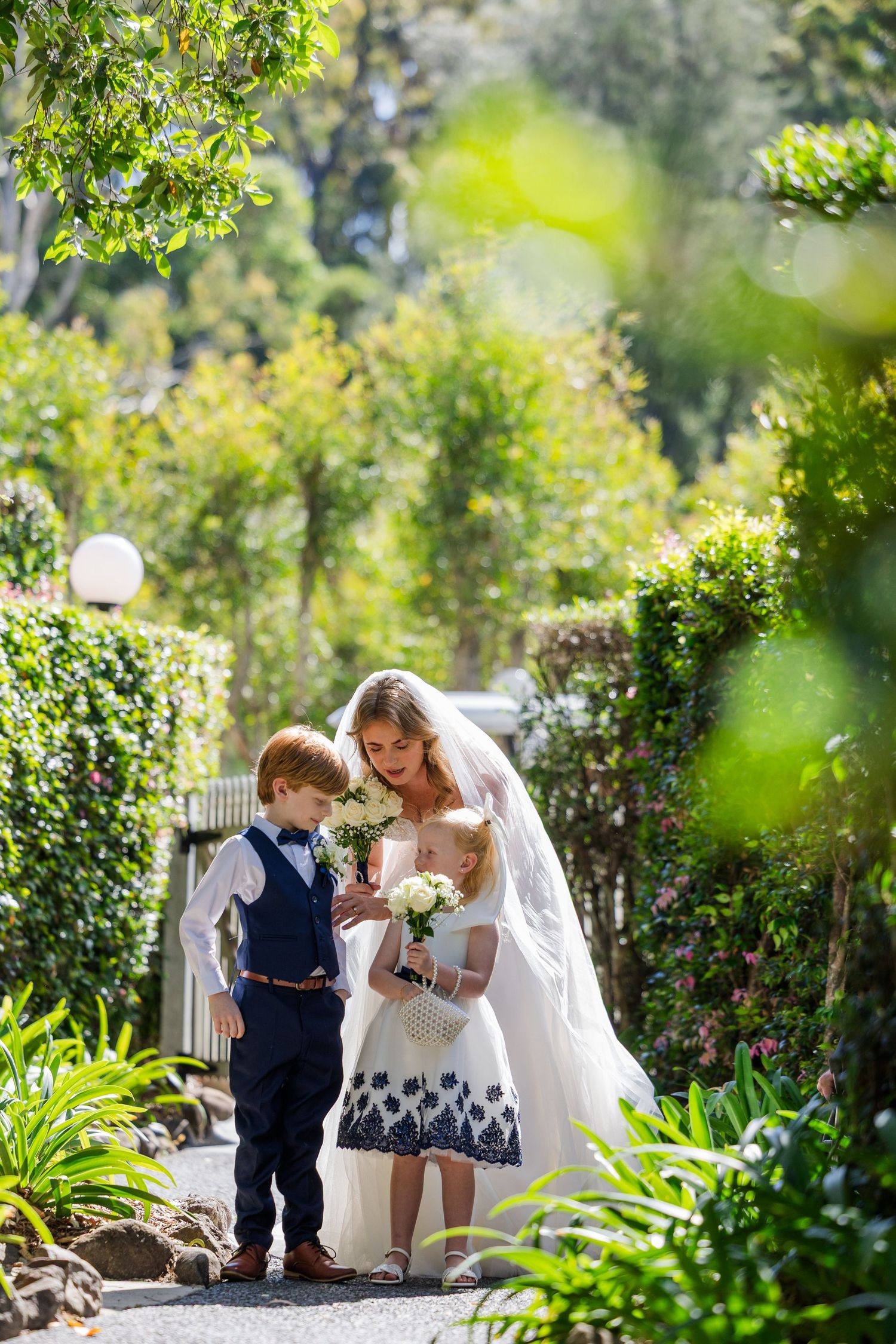 A bride in white shares a tender moment with a young child in a garden setting surrounded by lush greenery.