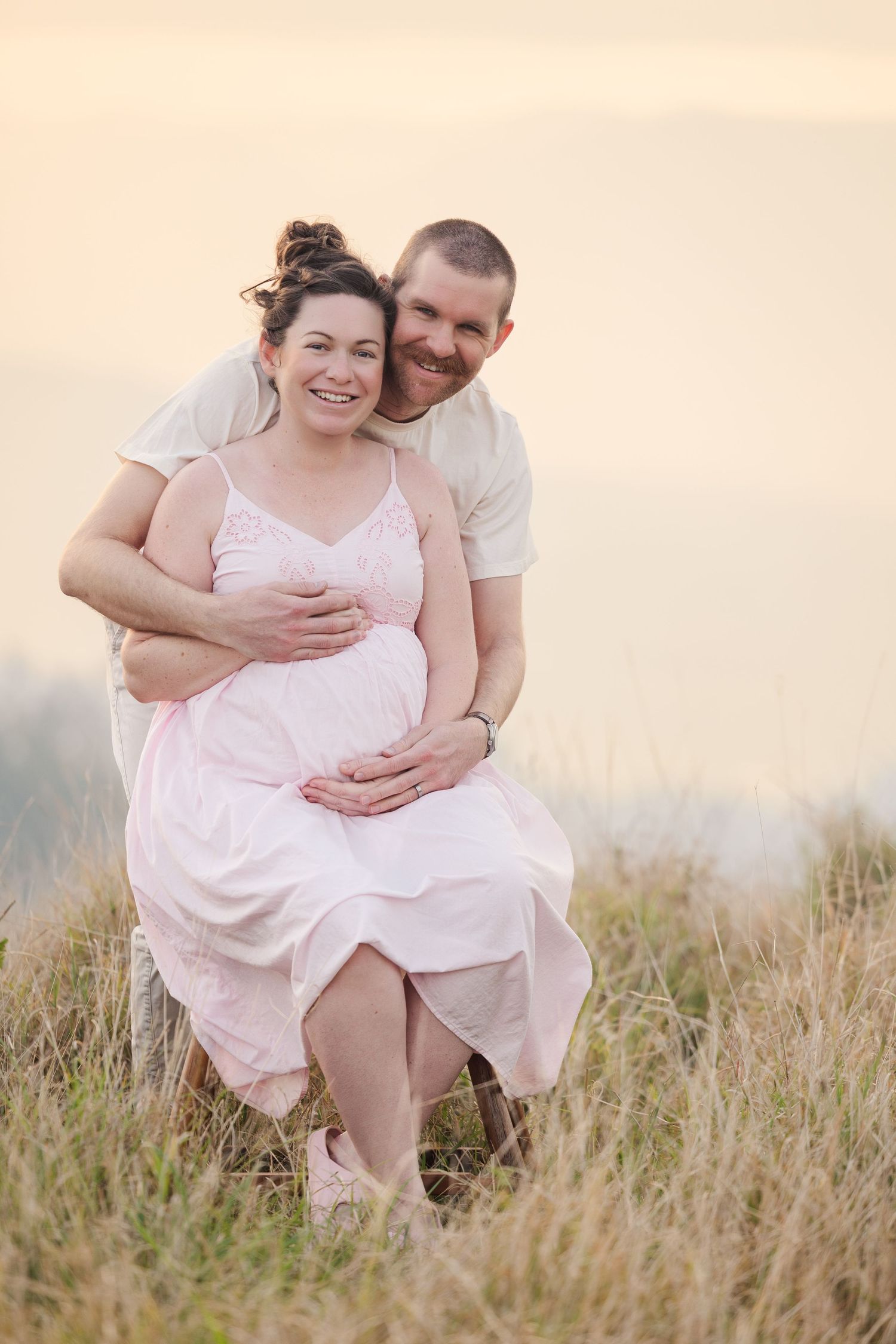 A couple embraces tenderly in a sunlit meadow wearing light colored clothing during a romantic outdoor photoshoot.