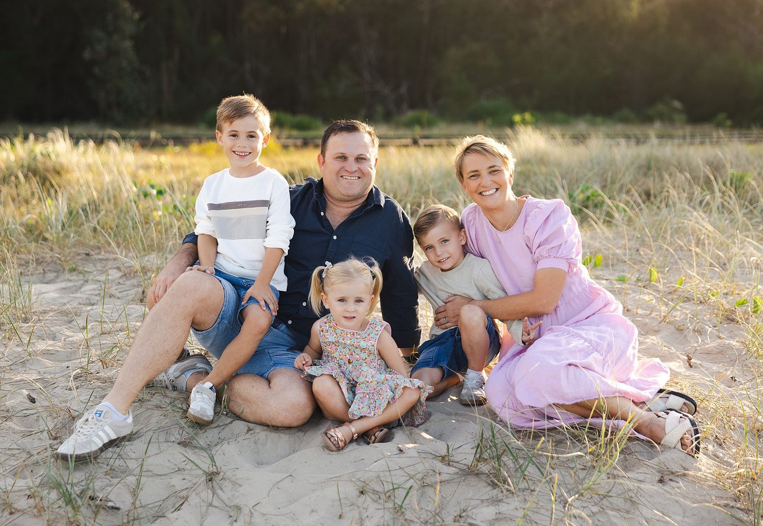 A group of five sits together in sunlit grass during golden hour, wearing casual outfits for an outdoor portrait.