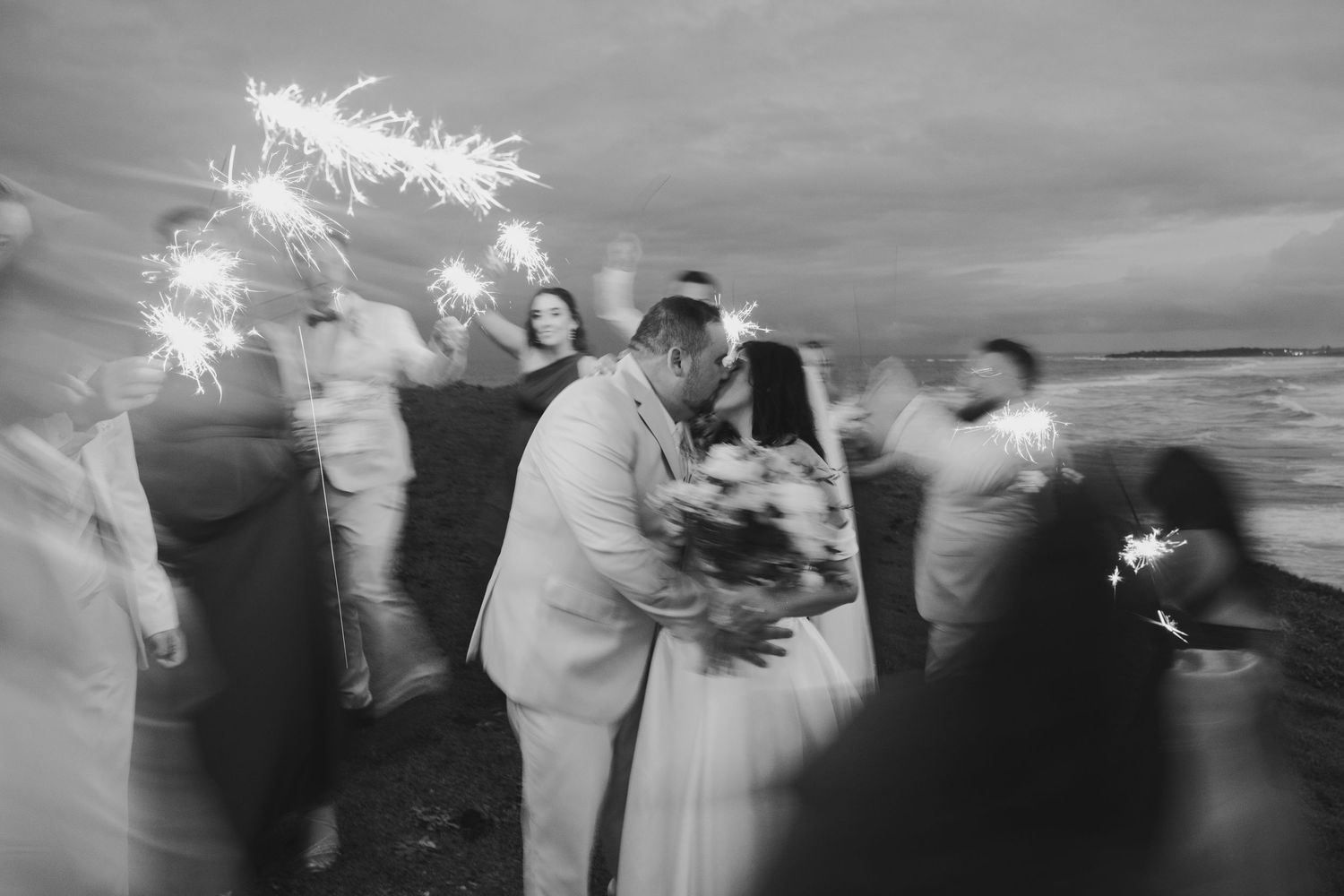 Black and white wedding photo of couple kissing surrounded by guests holding sparklers on a beach at dusk.