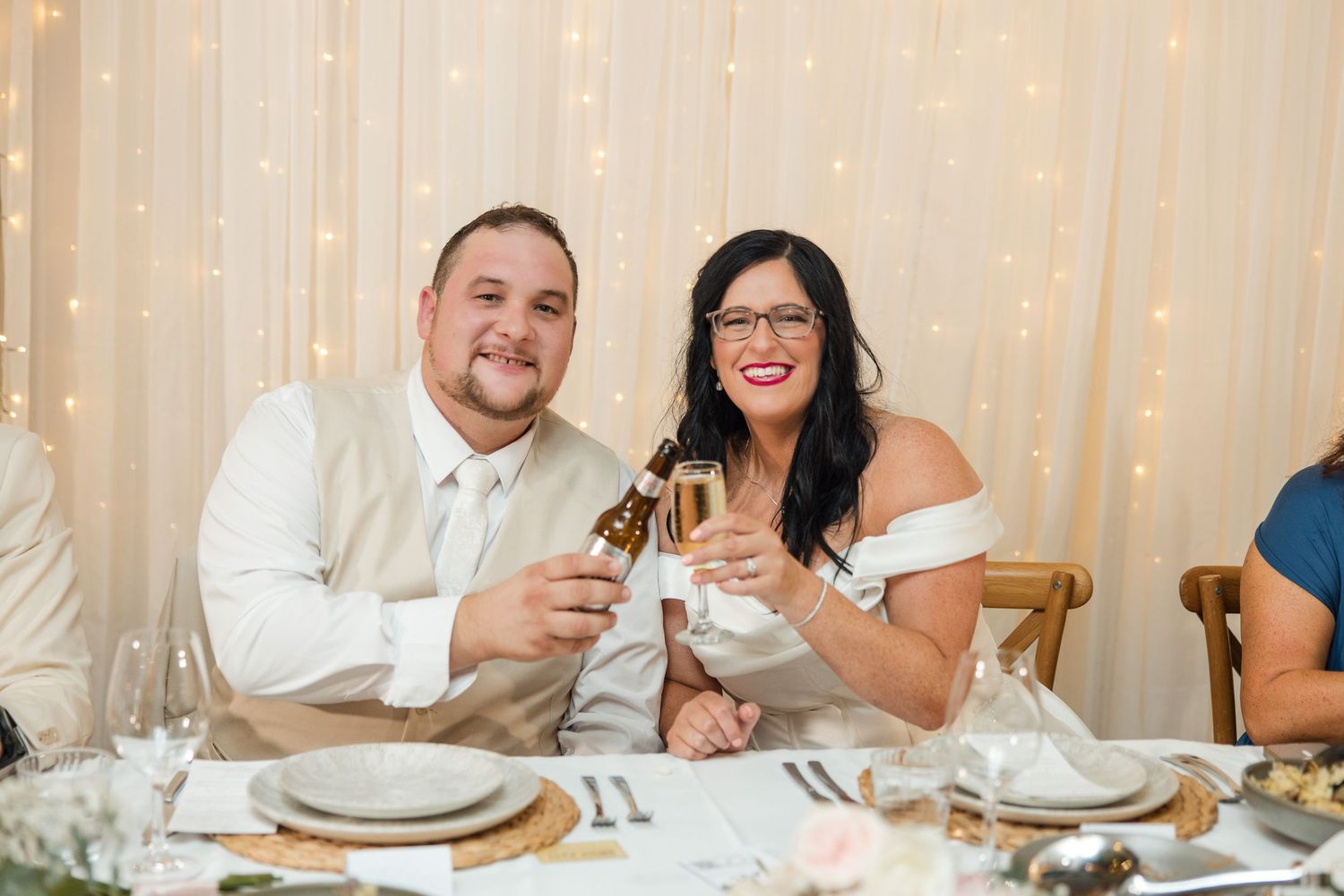 Bride and groom toasting with champagne and beer at wedding reception table with fairy lights backdrop.