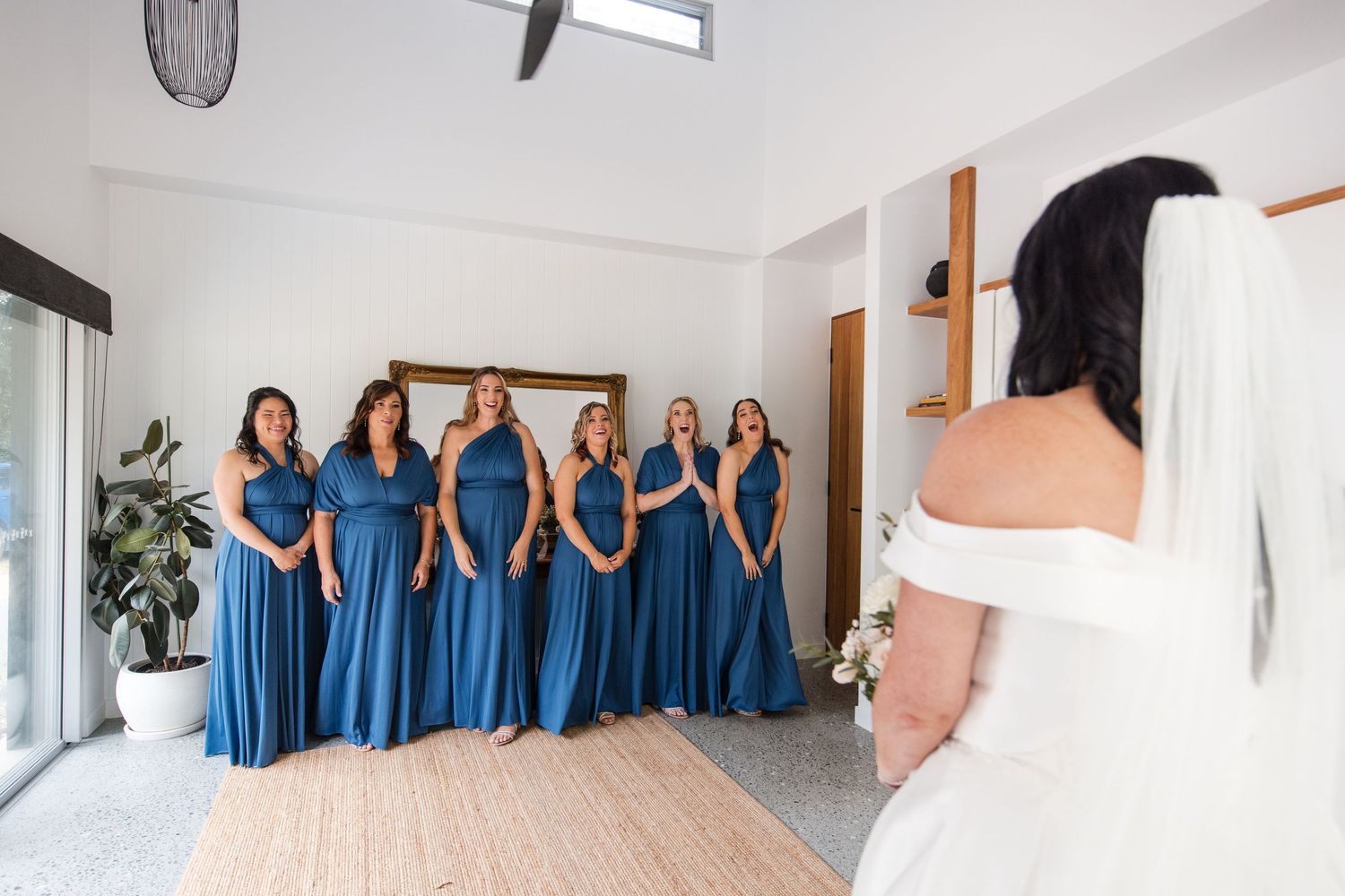 Bride in white gown reveals herself to bridesmaids in blue dresses during wedding morning preparations.