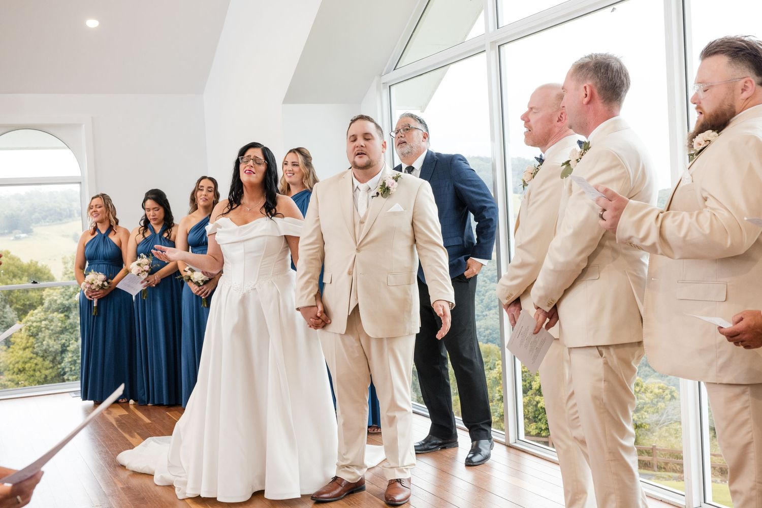Bride and groom stand with wedding party in blue dresses and tan suits before large windows with scenic views.