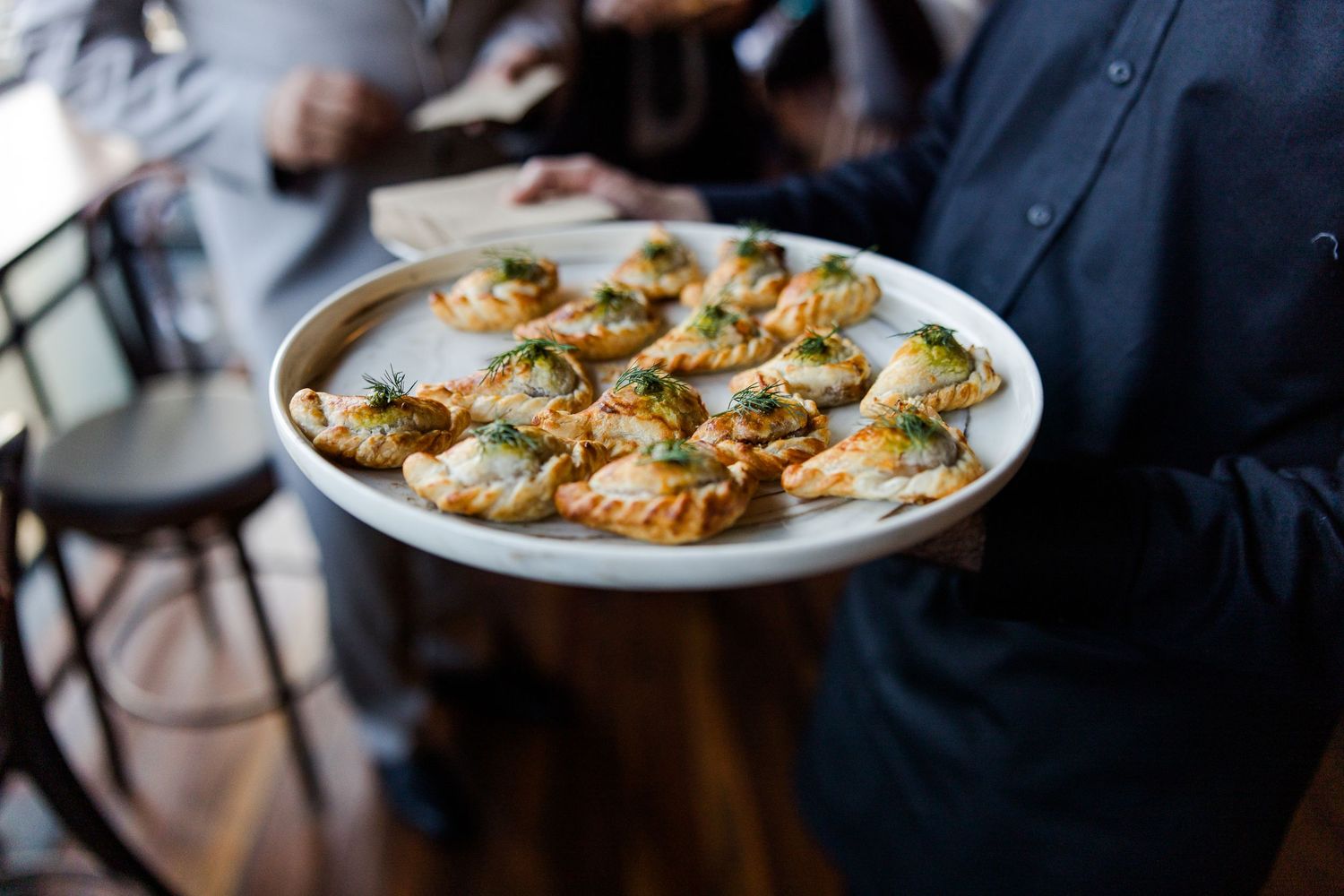 Waiter holding a round tray of elegant appetizers garnished with herbs at a catered event.