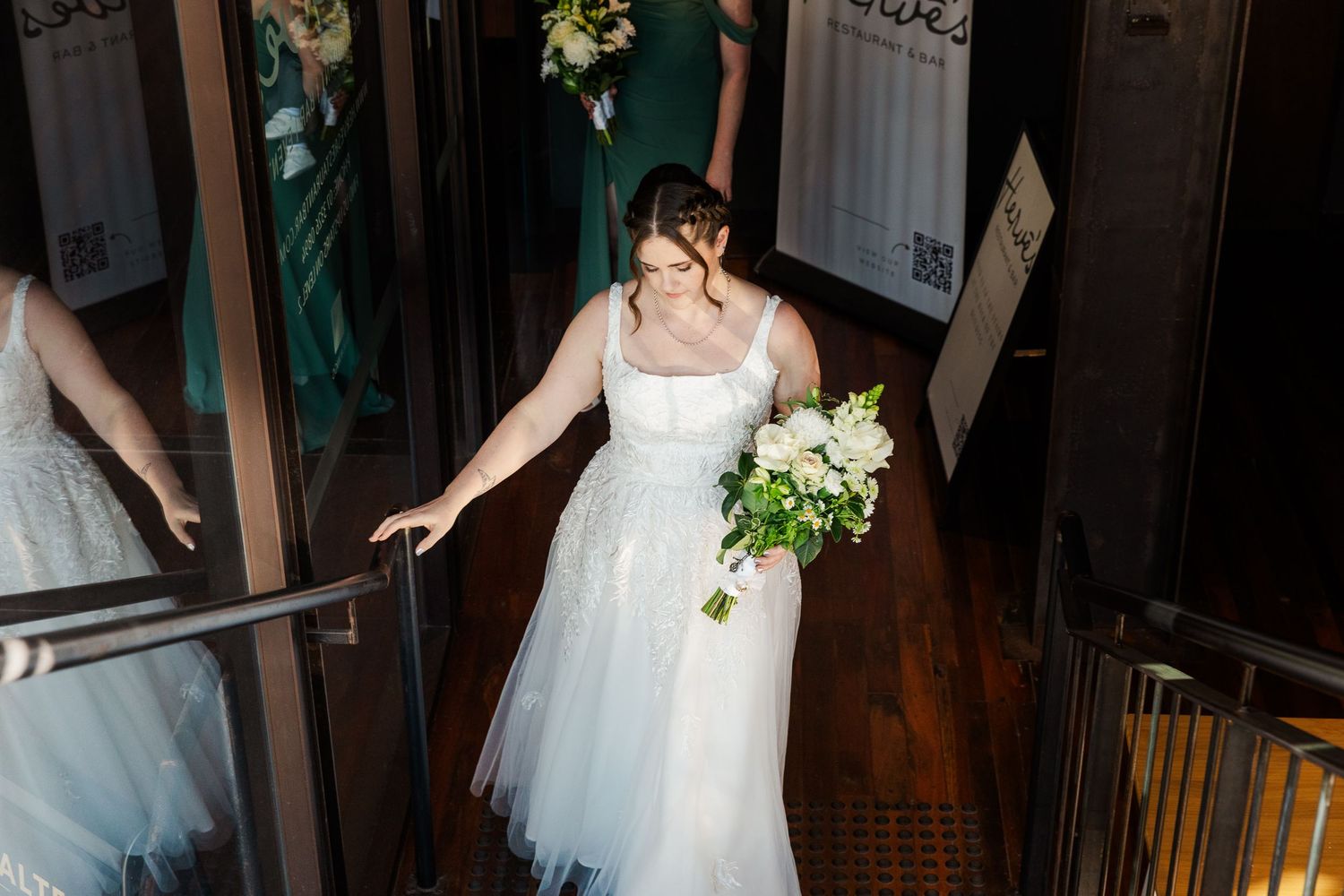 Bride in white tulle gown holding white bouquet descends staircase with green-dressed bridesmaids reflected in mirror.
