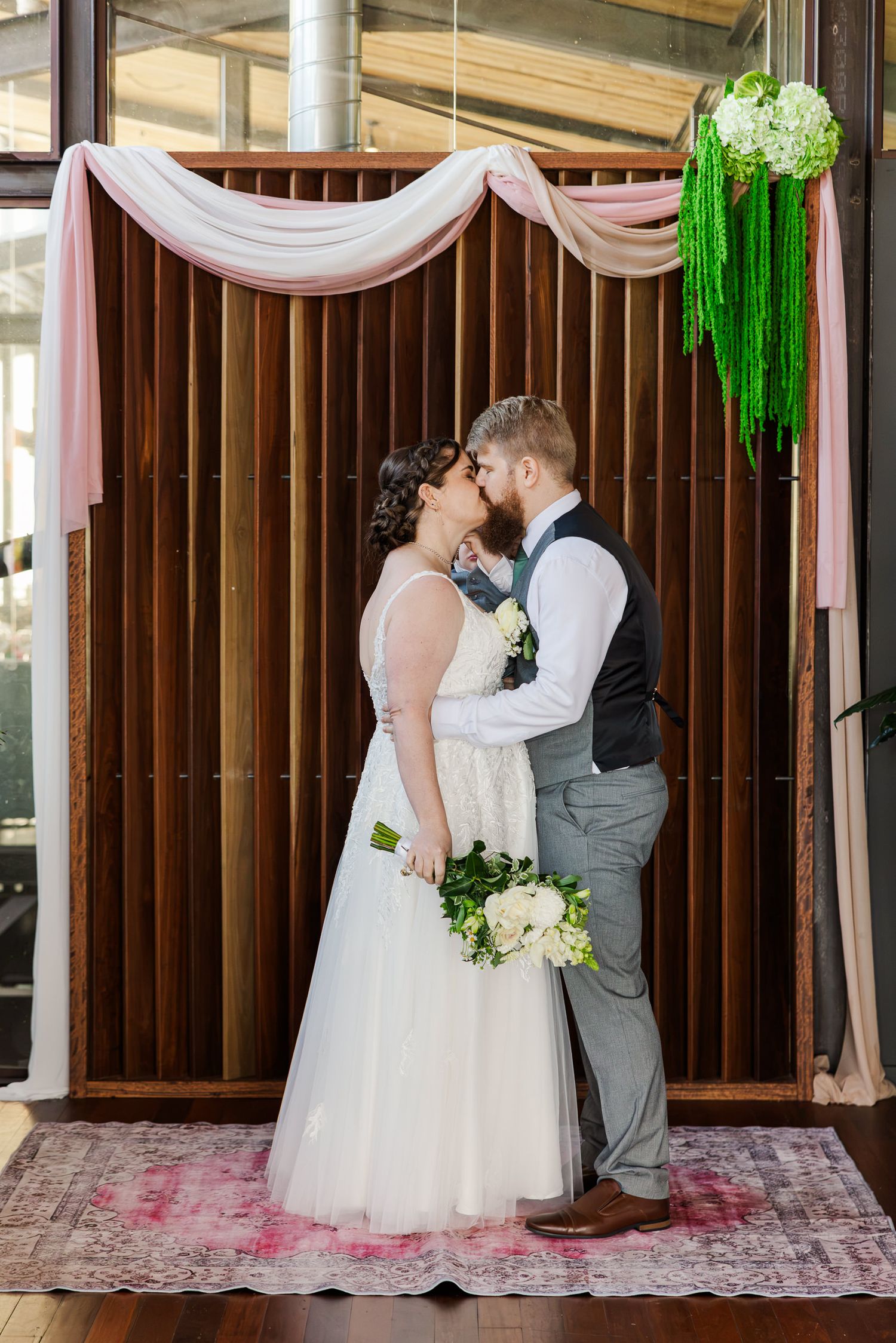Bride and groom share a kiss at the altar, decorated with pink draping and green floral accents, bride holding white bouquet.