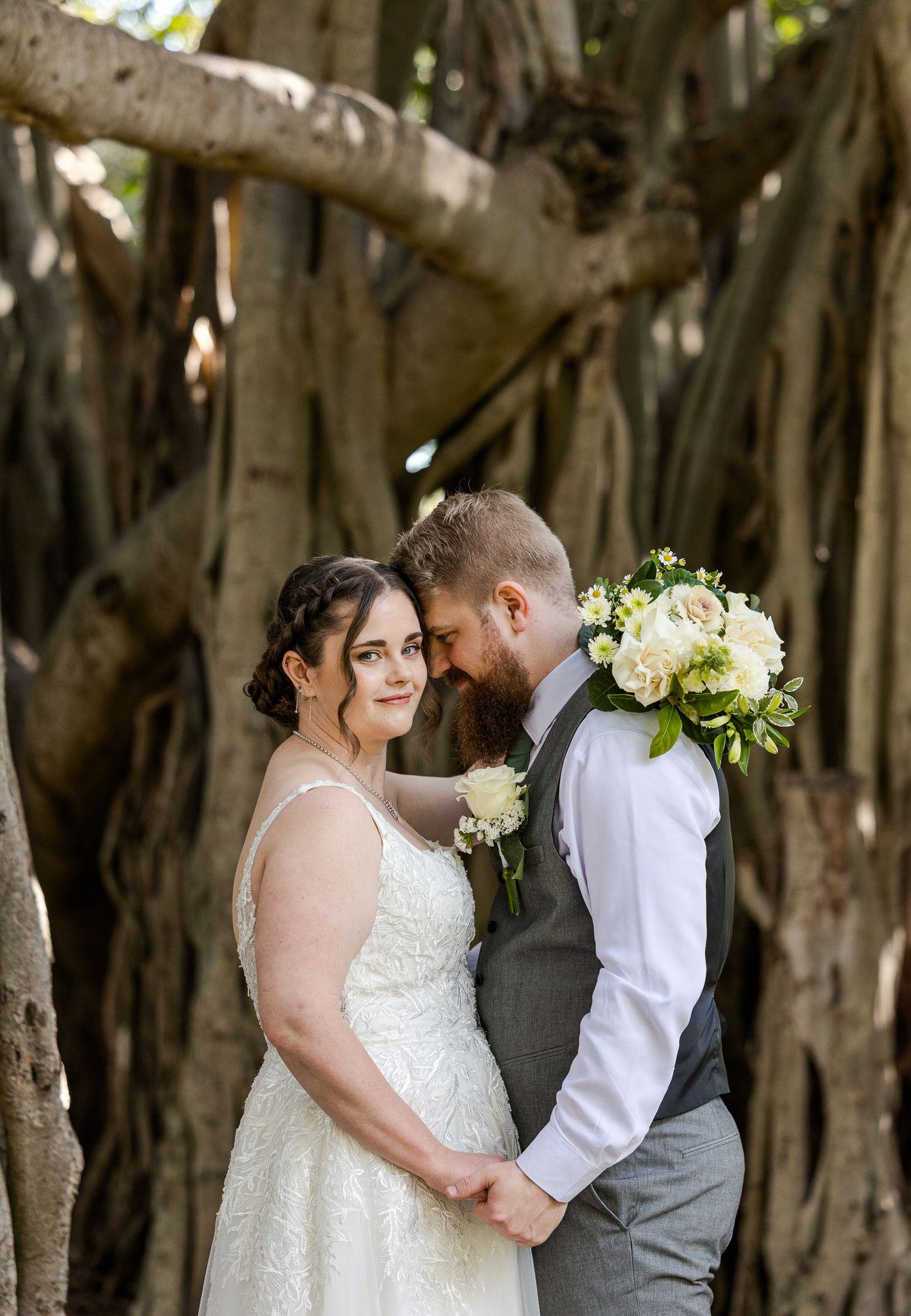 Bride and groom hold hands and touch foreheads under a large banyan tree, bride holds white floral bouquet.