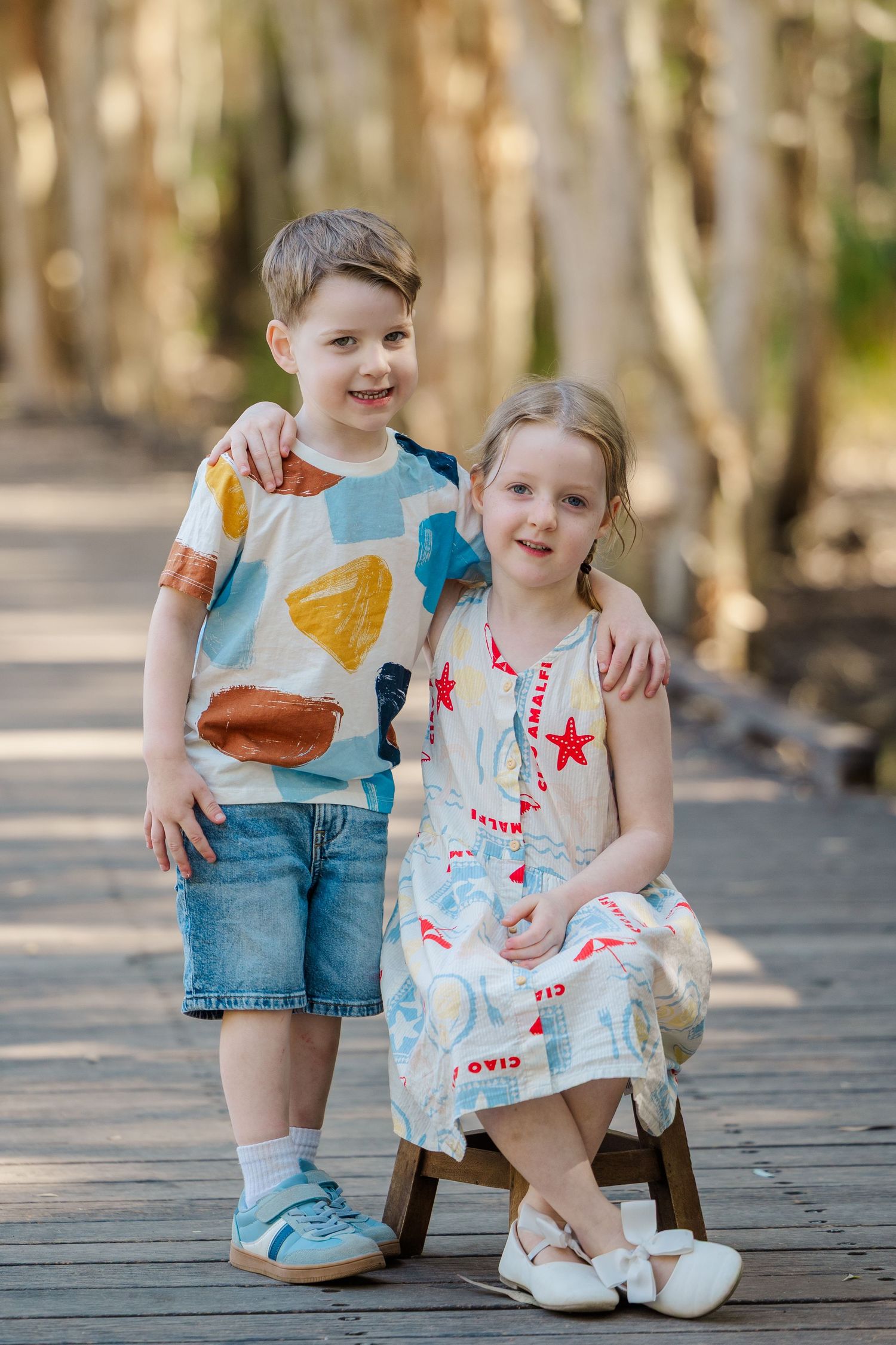 Two children sit together on a wooden boardwalk wearing coordinated summer outfits in blue and white.